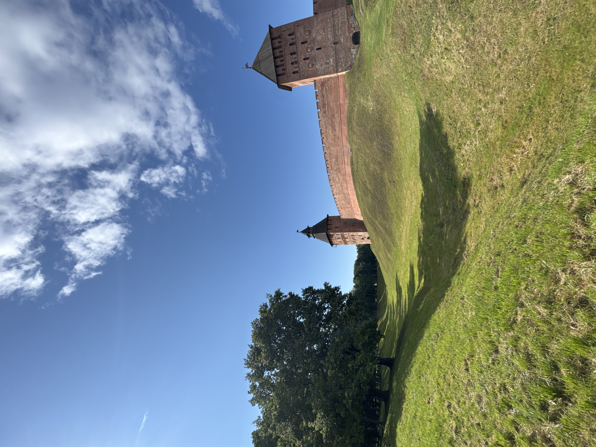 The image shows a section of the Novgorod Kremlin in Veliky Novgorod, Russia, on a clear, sunny day. The foreground features a grassy, undulating landscape, possibly a former moat, with the slopes covered in vibrant green grass. A large, mature tree with dense foliage stands to the left, casting a shadow on the grass. In the background, the red brick walls of the Kremlin rise majestically, topped with battlements. Two distinct towers are visible, one more prominent on the right with a wooden, conical roof and a small flag, and another further to the left, also with a dark, pointed roof. The sky is a brilliant blue, dotted with scattered white clouds. The scene is tranquil and evokes a sense of historical grandeur. There are no people visible in the image.
