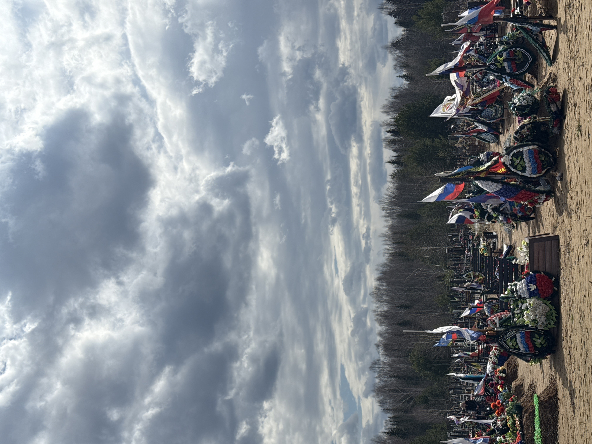 The image shows a section of the Southern Cemetery in Perm, Russia. Rows of graves are adorned with wreaths and Russian flags. The sky is overcast with heavy clouds. Bare trees line the background.