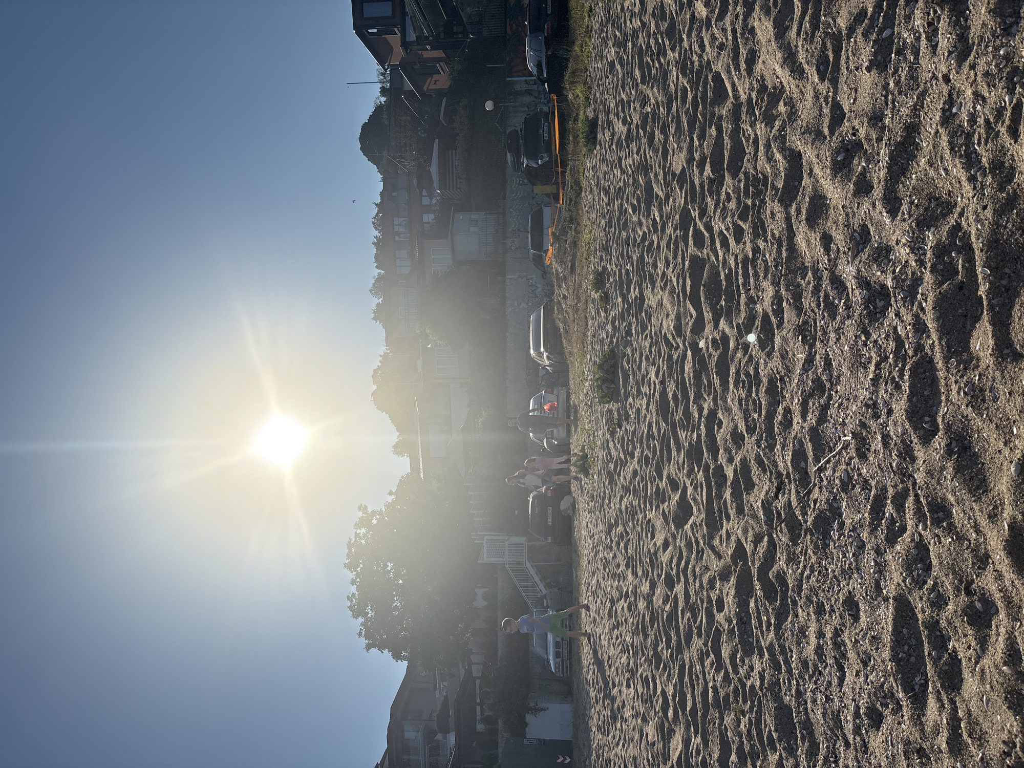 The image depicts a sun-drenched beach scene with a hillside covered in buildings in the background. The sun is bright and positioned high in the sky, suggesting it is midday and the weather is clear and sunny. The foreground is dominated by the textured sand of the beach, with ripples and some scattered shells visible.
On the hillside, a cluster of buildings, likely houses or apartments, are visible. Some trees and greenery are interspersed with the structures. A few cars are parked on a road or pathway leading up the hill, and several people are gathered near the cars, appearing to be socializing or perhaps preparing for an activity. One individual, a young boy in blue shorts and a green shirt, is seen in the middle ground, standing on the sand and looking towards the left of the frame.
The overall mood of the image is relaxed and summery. The bright sunlight and the presence of people on the beach suggest a leisurely day. There is no visible text in the image. The exact location is unknown.