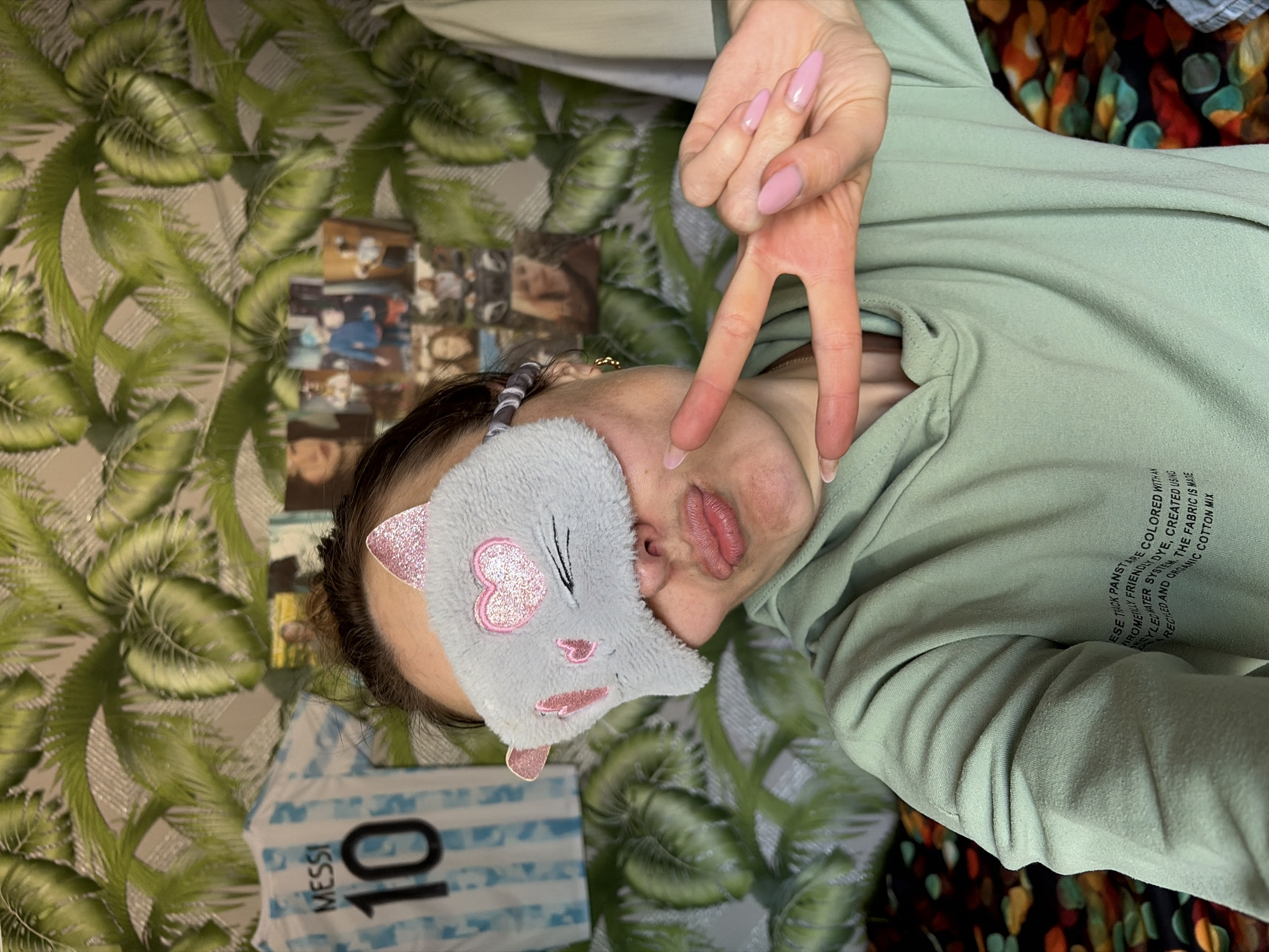 A woman wearing a light green sweatshirt and a gray sleep mask shaped like a cat with pink glitter accents is lying on her back. Her hand is raised, making a peace sign. She has light pink nail polish. Her lips are slightly parted. A soccer jersey with "MESSI" and the number "10" is partially visible. The background includes a wall with a palm leaf pattern and a collage of photos. Text is visible on the sleeve of the sweatshirt, including "TRACK PANSTRE COLORED WITH AN" and "RECILED AND ORGANIC COTTON MIX". The location is Nizhnii Vishur, Russia.