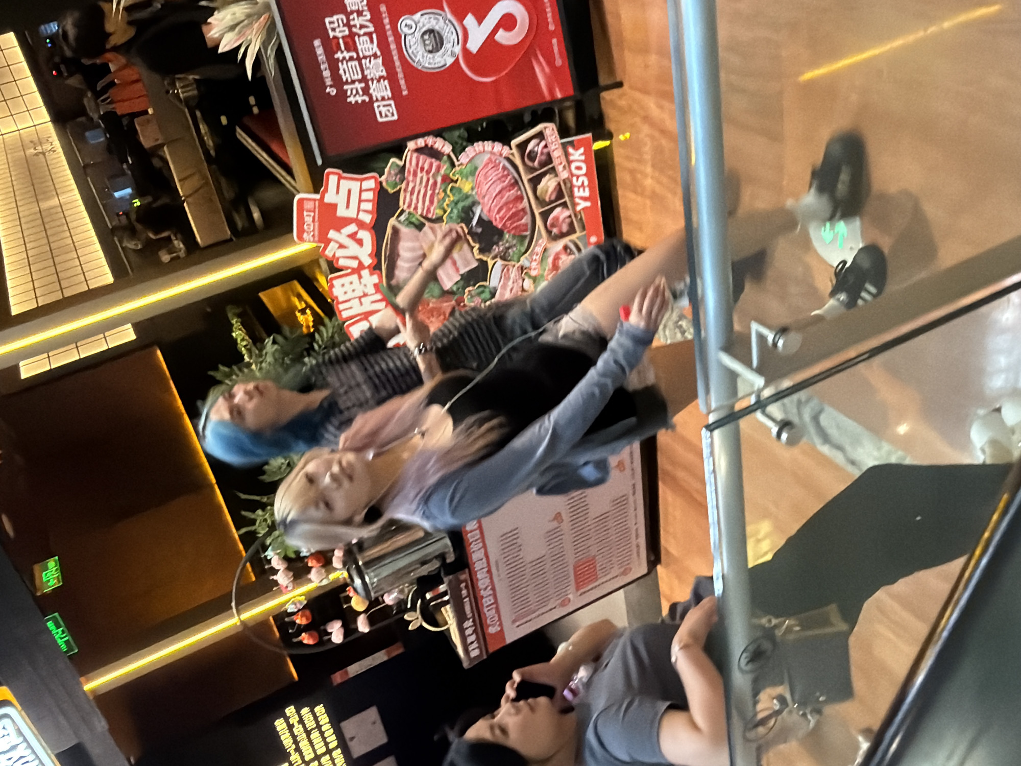In a commercial setting, likely a shopping mall in Yuzhong District, China, a group of individuals is captured on an escalator. The scene is illuminated by ambient lighting, with neon accents and signs visible. In the foreground, a person with dark hair is seen from the chest down, wearing a grey t-shirt and holding a mobile phone to their ear. Beside them, a person with blonde and pink-tinted hair, wearing a light blue cardigan over a black top and shorts, is looking towards the camera. They are adorned with necklaces. Further back, a person with blue hair, dressed in a striped shirt and grey pants, is partially visible, looking to the right. A large red advertisement sign featuring images of raw meat and Chinese text is prominently displayed, along with smaller signs and promotional materials. A stainless steel dispenser is also present. The escalator's glass railing reflects the surrounding environment and the people present.