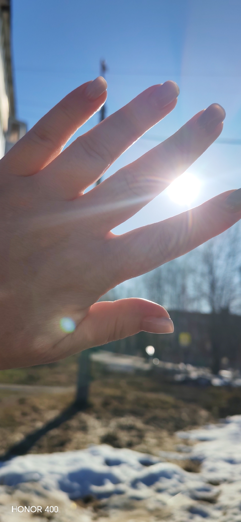 The image shows a person's hand held up towards a bright sun. The fingers are spread open, and the sunlight filters through them. The background is blurred, showing a landscape with patches of snow, some buildings, and bare trees. The photo was taken with an HONOR 400 phone. The exact location within the city of Noviy, Russia is not discernible from the image.