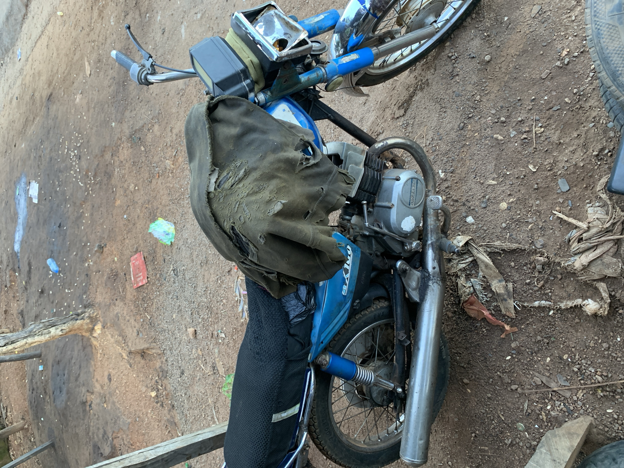 A weathered blue and white motorcycle is parked on a dirt road in Kabba, Nigeria. The motorcycle is covered with a torn, dark green fabric that is ripped in several places. The seat of the motorcycle is covered with a black mesh material. The front of the motorcycle has a chrome headlight and blue front suspension. The rear of the motorcycle has a silver exhaust pipe and a blue shock absorber. The ground around the motorcycle is covered in dirt, gravel, and scattered debris, including plastic bottles, paper, and what appears to be dried organic matter. In the background, a weathered wooden post and a dead tree trunk are visible. The environment appears unkempt and rural.