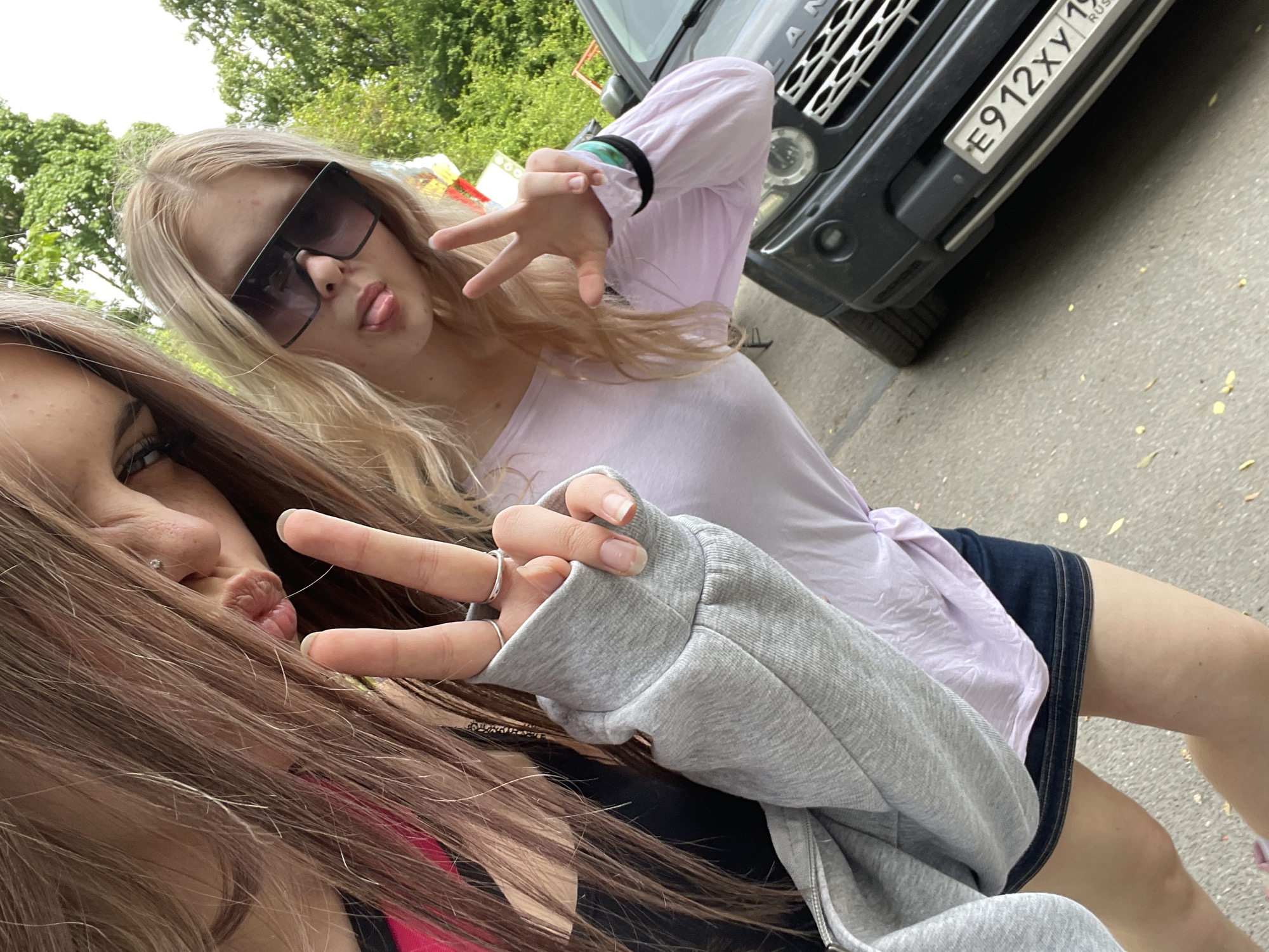 In Lednevo, Russia, two young women pose for a close-up outdoor photograph. The woman on the left, with long light brown hair and a nose piercing, wears a black top under a gray zippered hoodie. She makes a peace sign with her left hand, wearing two rings, and extends her tongue. The woman on the right, with long blonde hair, wears oversized dark sunglasses, a light purple long-sleeved top, and a dark denim skirt. She mirrors the pose, making a peace sign with her right hand, which is adorned with a green and black wristband, and extends her tongue. Behind them, a dark-colored SUV, identifiable by the partial text 'LAN' on its grille, is parked, displaying a Russian license plate reading 'E912XY 19 RUS'. The ground is a paved surface with scattered yellow leaves, and green tree foliage is visible in the upper background.