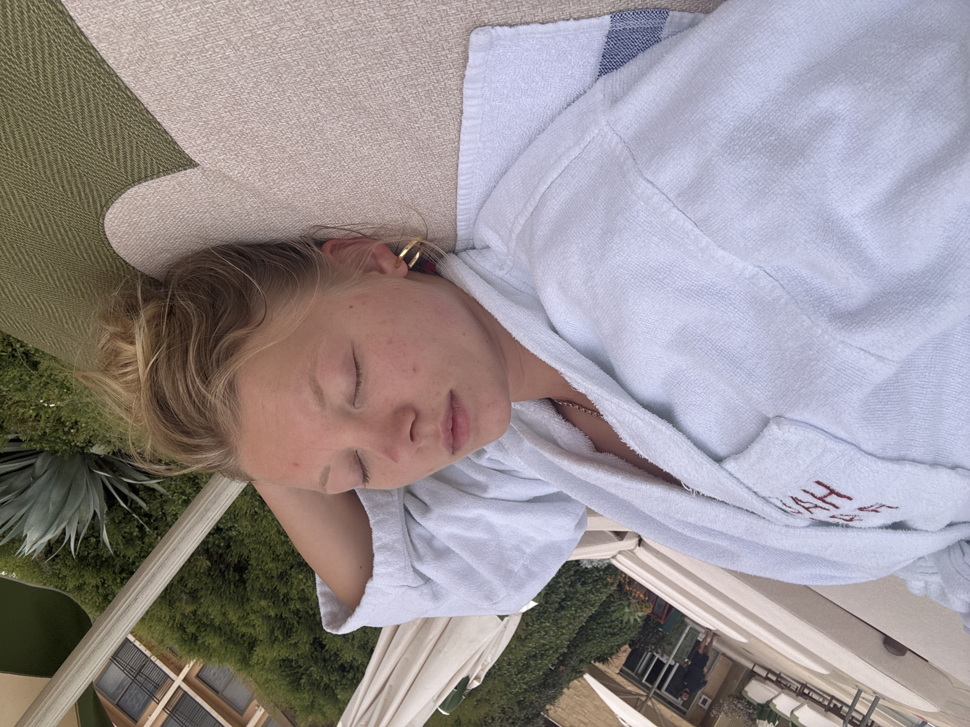 A young person is sleeping on a lounge chair outdoors. The person is wearing a white bathrobe with red lettering on the pocket area. A gold hoop earring is visible in their left ear. The lounge chair is tan and green. Lush green foliage and a building are visible in the background. Based on the lettering "Herzliya" on the robe, the location is likely Herzliya, Israel.