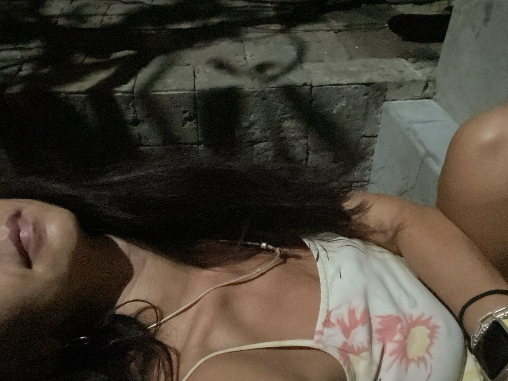 This low-light image offers a close-up, angled view of a person, likely a woman, from the neck to the mid-chest and shoulder, reclining or resting. Her dark hair cascades over her shoulder, partially obscuring her face, though her lightly parted lips are visible. She wears a light-colored top, possibly a sundress or camisole, featuring a floral pattern with pink and yellow blooms. Around her neck is a thin, light-colored necklace with small bead-like elements, and a delicate white cord rests along her collarbone, possibly an earphone wire. On her left wrist, she wears a black elastic band and a silver-toned bracelet or watch with a dark, square face.
The setting appears to be outdoors at night or in very dim light, evidenced by the darkness and pronounced shadows. Intricate dark shadows, resembling foliage or branches, are cast upon a textured paved surface, likely stone or concrete blocks. Portions of a light grey stone structure, possibly a bench or low wall, are also visible in the background. The overall mood suggests a moment of calm and relaxation. The context provided indicates this location is Kuta Selatan, Indonesia. No discernible text is present in the media file.