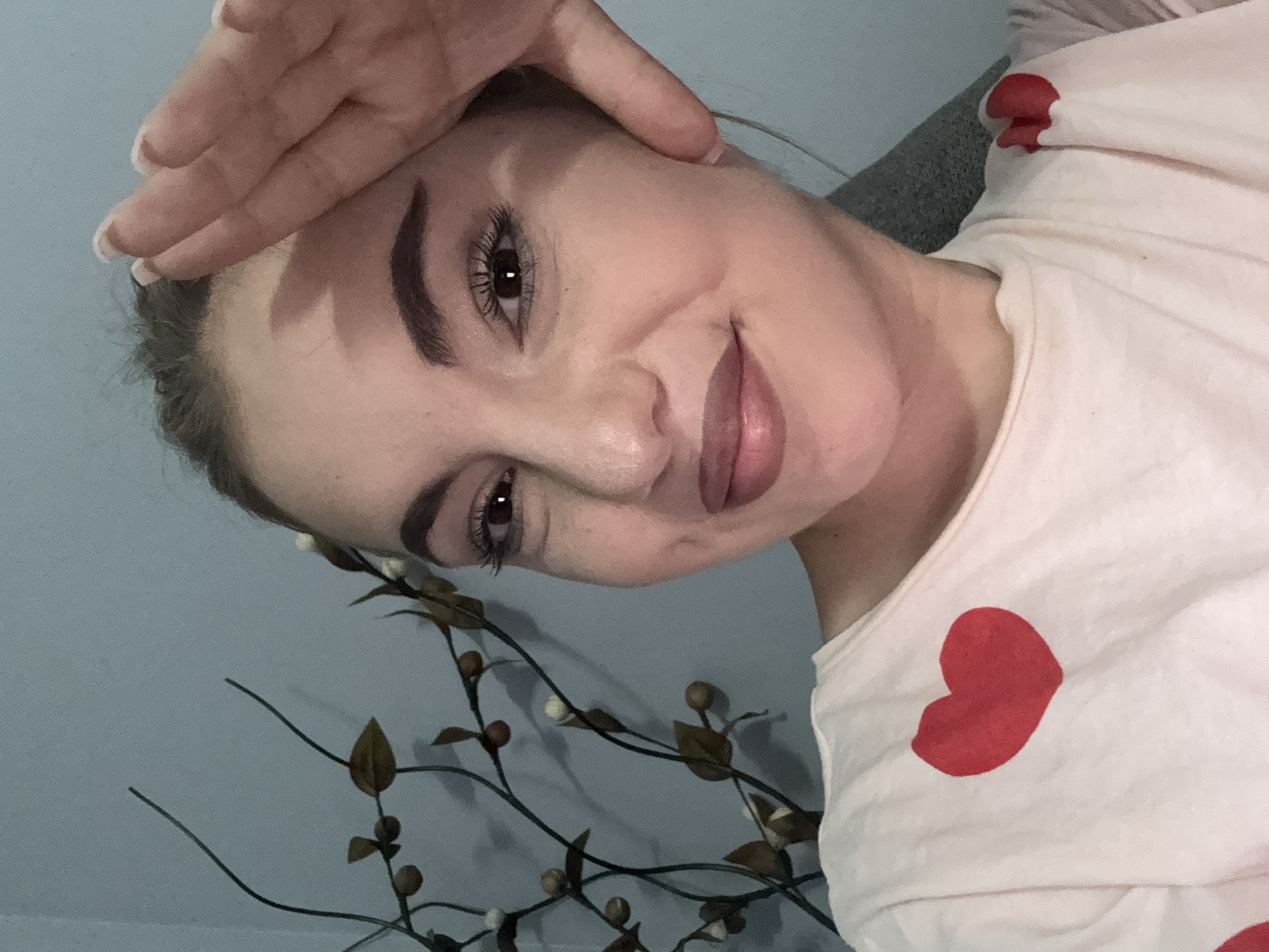 A young adult female is depicted in a close-up, frontal portrait, likely a selfie, within an indoor setting. She has dark hair pulled back and dark brown eyes, with visible makeup including eyeliner, mascara, and a brownish-pink lip product. A subtle smile is present, and a dimple is visible on her right cheek. Her right hand is raised, with fingers slightly spread, the palm partially covering her upper forehead and hairline, casting a shadow across the upper portion of her face. Her nails are manicured with white tips. She is wearing a light-colored t-shirt adorned with multiple red heart designs; one prominent heart is on her left chest area, and a partial heart is visible on her right shoulder. The background consists of a plain light blue wall. To the left of the subject, a decorative element composed of dark, thin branches with small brown leaves and light-colored spherical buds casts shadows on the wall. The lighting appears direct, creating distinct shadows. The image provides no discernible visual information to indicate its specific geographical location within Sofia, Bulgaria, or any other city.