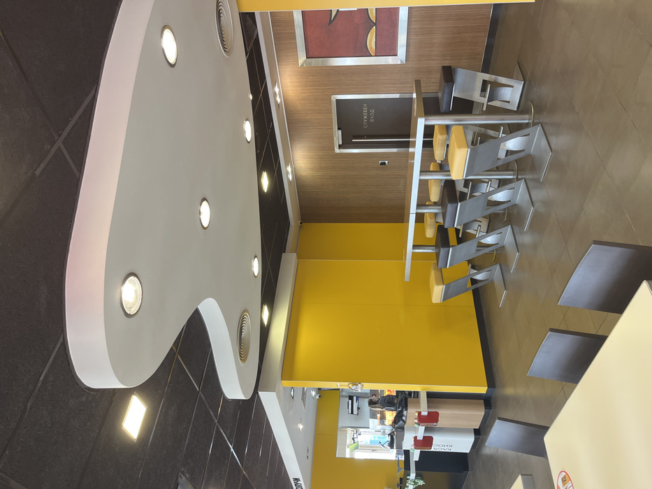 The interior of a fast-food restaurant in Hadjidimitrovo, Bulgaria, featuring a white, abstract, curved ceiling with recessed lighting, a yellow wall, a brown wood-paneled wall, and a small, empty seating area with a long, narrow table and six stools with yellow cushions. The wall on the right has a sign that reads "SERVICE ENTRANCE." A single, female employee is visible in the background, near a counter with red signage and a small, potted plant. The floor is tiled in a neutral color. The restaurant is clean and well-lit. The scene is quiet and empty, with no customers present.