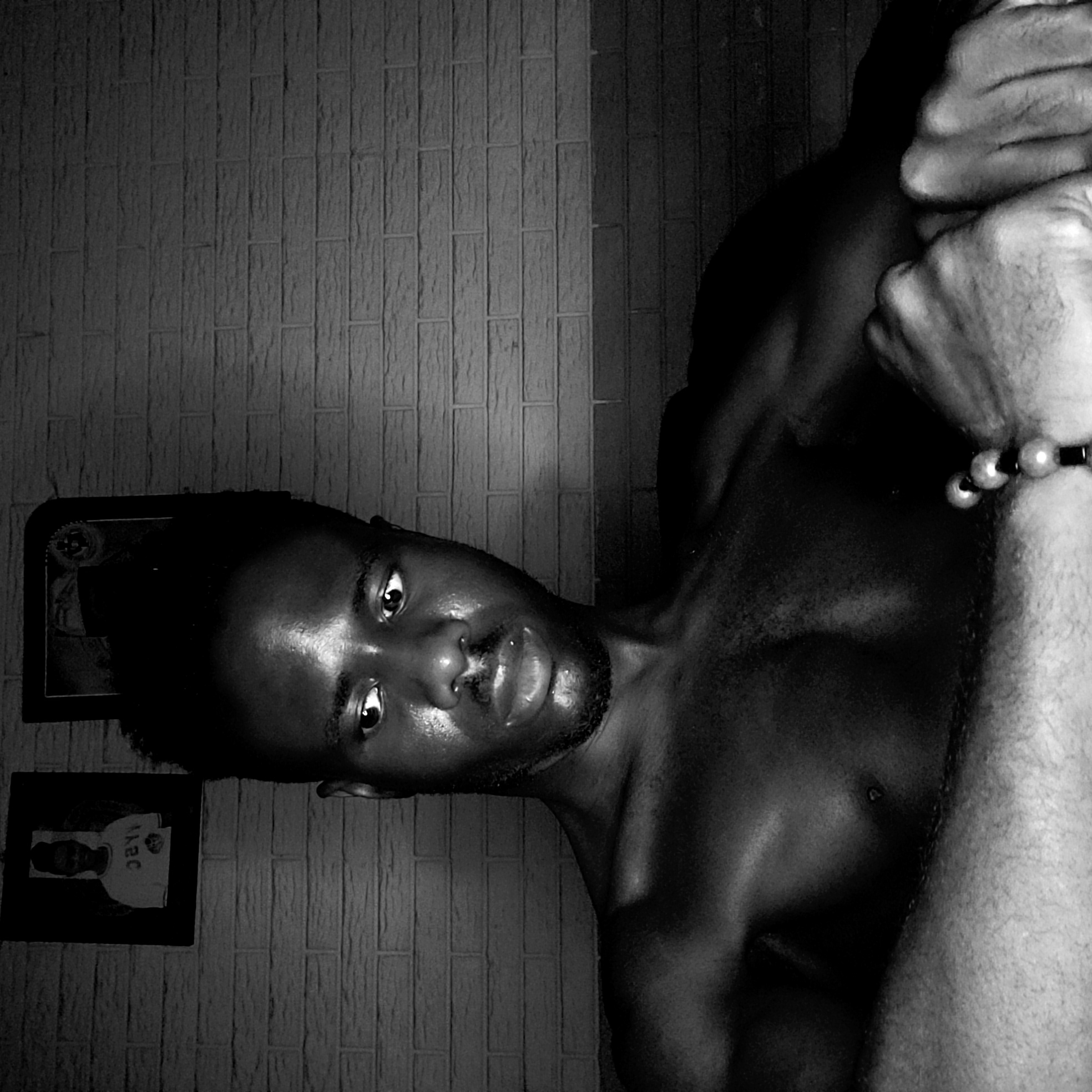 This black and white portrait features a young, dark-skinned man in an indoor setting, likely in Narayi, Nigeria, as suggested by the location context. The image is composed with high contrast, highlighting the man's features and creating dramatic shadows.
The man, appearing shirtless, is positioned with his head slightly tilted and his gaze directed intently towards the camera. His skin, particularly on his face, chest, and shoulders, has a glossy, almost oily sheen, catching the light. He has short dark hair, a beard, and a mustache. On his right wrist, he wears a bracelet made of light-colored beads interspersed with dark spacers. His right arm is bent and appears clenched, with his hand visible in the upper right portion of the frame.
Behind the man is a wall with a distinct, textured pattern resembling rectangular bricks or tiles. Two framed pictures are hanging on the wall to the left of the man. The upper frame is partially visible, showing a dark-toned image. The lower frame contains a picture of a person wearing a white t-shirt, on which reversed text is faintly visible, appearing to spell out "A2C" or similar characters.
The strong, possibly artificial, lighting creates deep shadows and accentuates the contours of his body, contributing to the dramatic and intense mood of the photograph. The man's direct stare and strong pose convey a sense of confidence and presence.