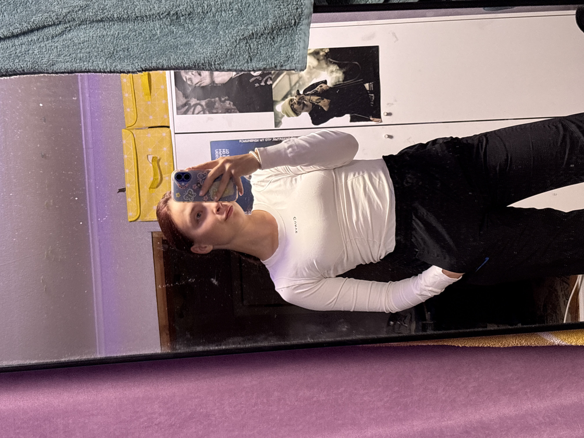 A mirror selfie taken indoors. The reflection shows a woman with short auburn hair wearing a long-sleeved white top and black sweatpants. She's holding a cell phone with a patterned case. In the background, there's a white cabinet with posters and yellow boxes. The surface beneath the mirror is a purple fabric. There is a green towel over the top of the mirror. The city is not discernible.