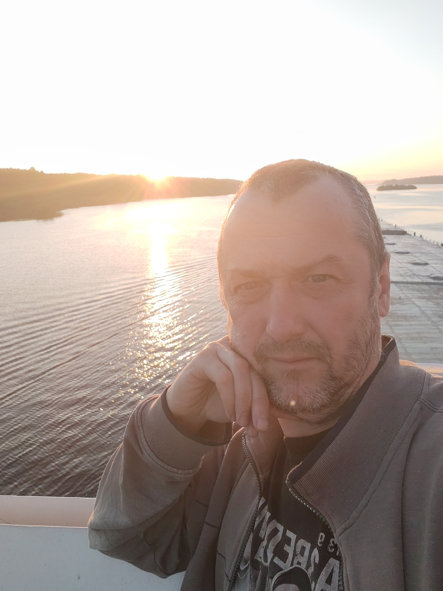 A medium shot shows a Caucasian man with a beard and short hair, wearing a casual jacket, resting his chin on his hand. The background reveals a landscape with water, trees, and a setting or rising sun. A structure resembling a pier or road is visible on the right side. The scene appears to be outdoors during the golden hour. There are no indications that this picture was taken in Sud'bitsy, Russia.