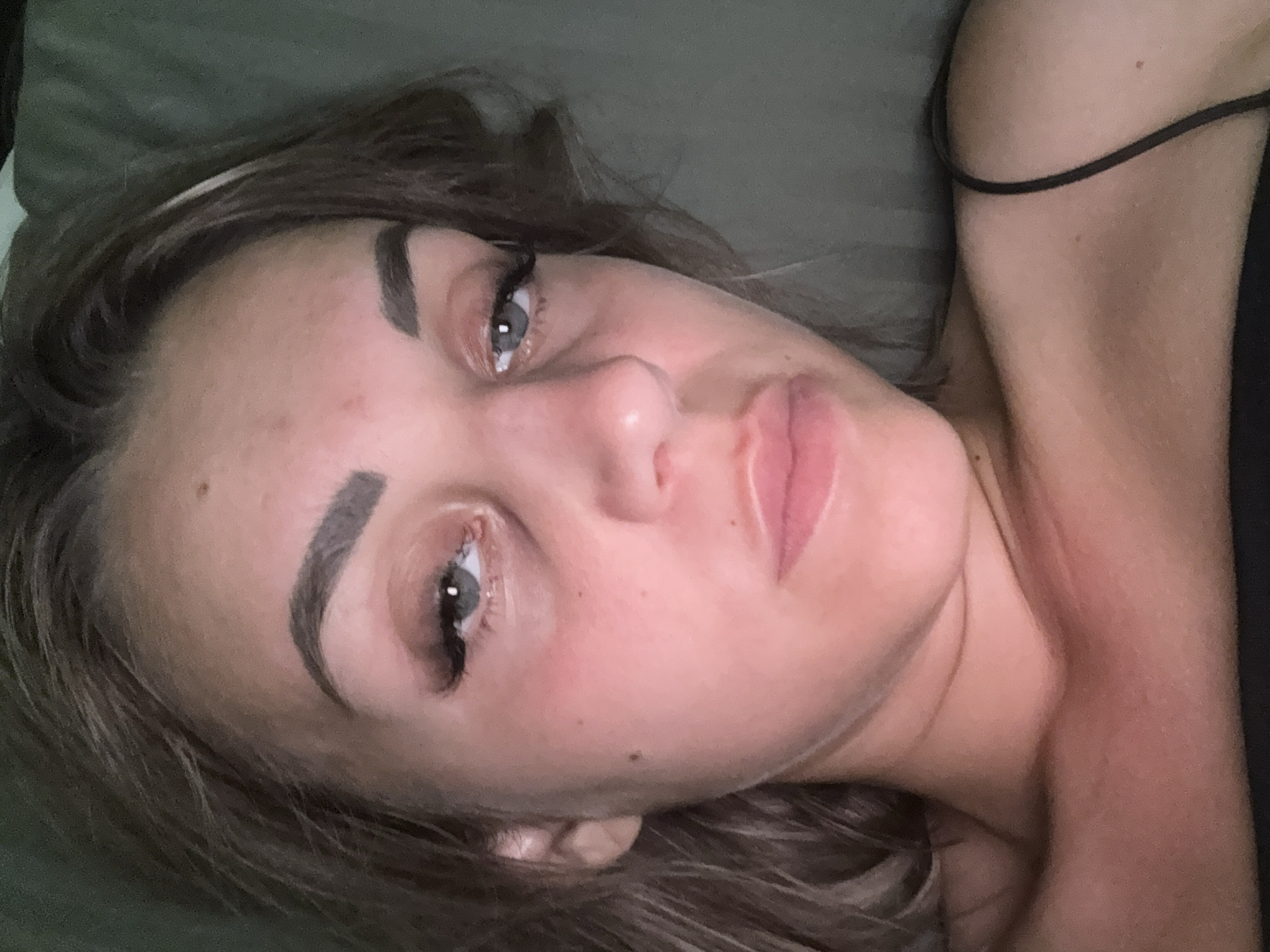 The image is a close-up portrait of a young woman lying down. She has light brown hair, prominent eyebrows, and is wearing false eyelashes. Her eyes are looking upwards, and her mouth is slightly open. Her skin appears to have a pinkish hue, possibly due to lighting or blush. She is wearing a black top with a thin strap visible on her right shoulder. The background is a dark, textured surface, likely a bed or couch. There is no discernible location in Yuzhno-Sakhalinsk, Russia, or any urban environment visible in the image. The scene is domestic and personal.