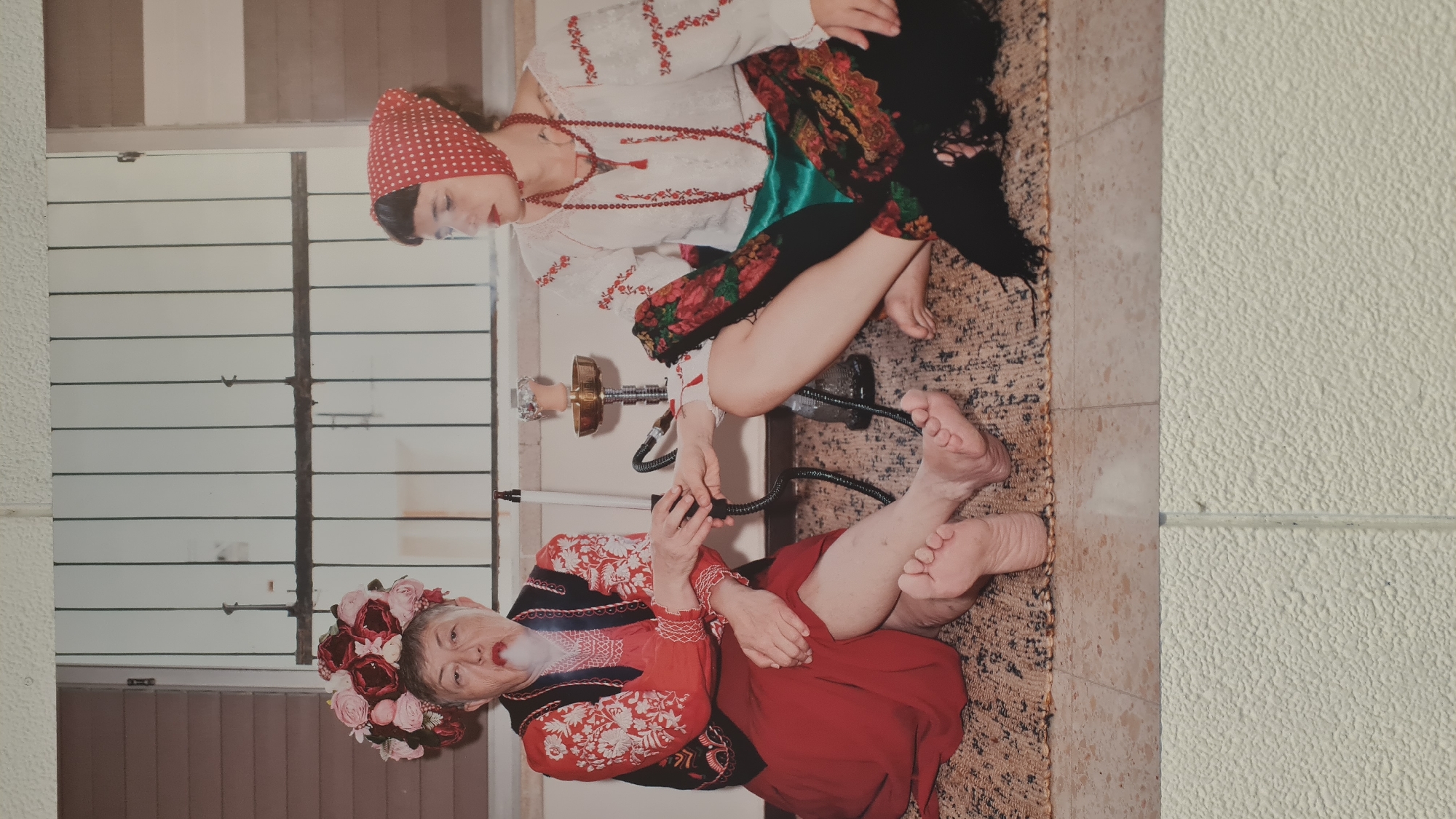 The image shows two women in traditional Eastern European clothing, likely folk costumes. One woman, dressed in a white embroidered shirt, red polka-dot headscarf, and a red beaded necklace, is sitting cross-legged. The other woman, who appears older, is wearing a red and black embroidered dress and a wreath of pink and red flowers on her head. They are sharing a hookah, with the older woman taking a puff from the hose. The environment suggests an interior setting with a window featuring horizontal louvers. The floor is tiled. The photograph may have been taken in Nahariyya, Israel, but no specific location can be discerned from the image itself.