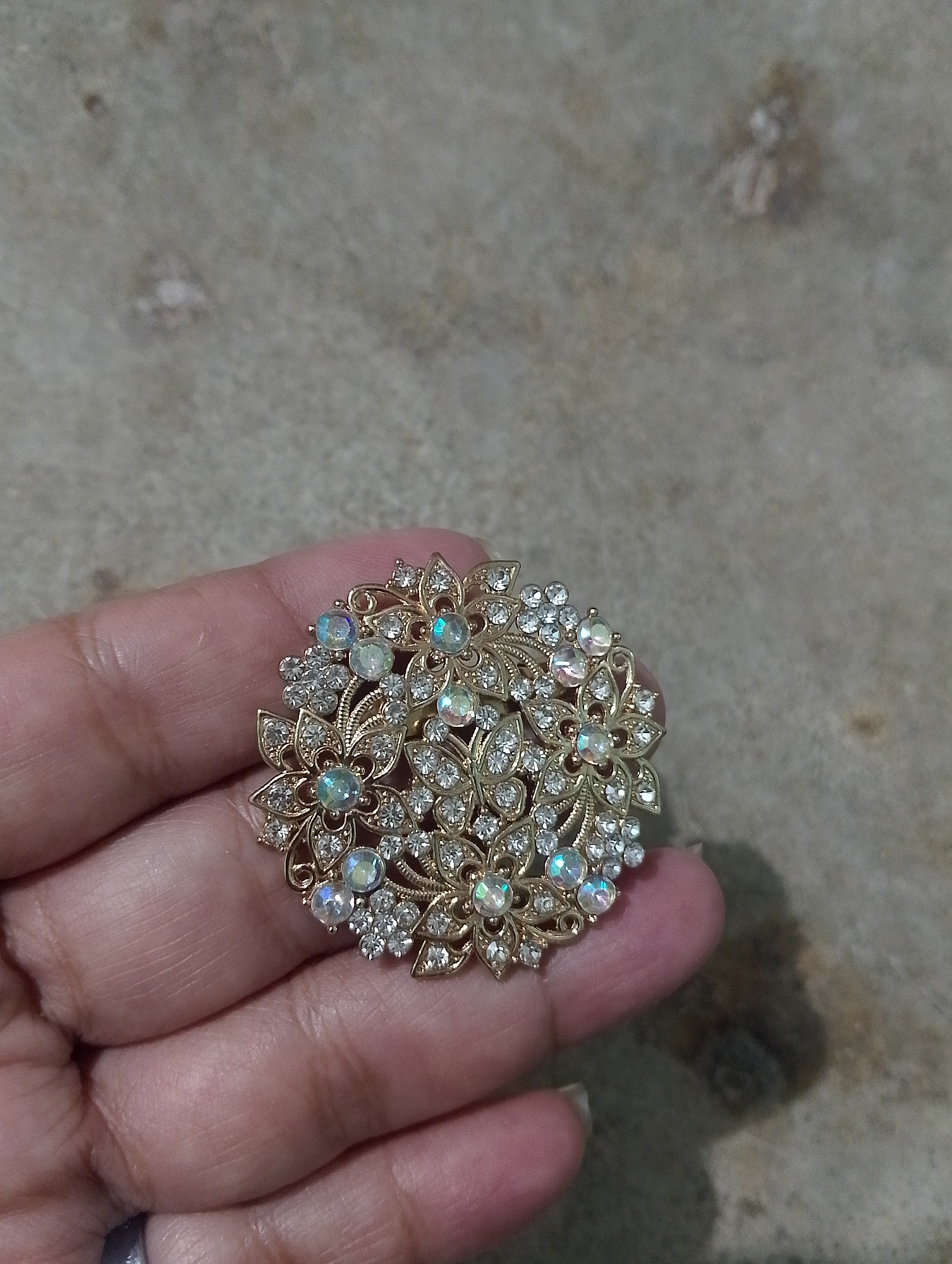 A gold-toned brooch with clear and iridescent rhinestones is held in a person's left hand, which rests on a mottled gray surface. The brooch is circular and features a floral design with intricate metalwork and a scattering of small stones around larger, multifaceted ones. The background surface appears to be a concrete or stone patio. The image provides no information about the city of Serbelawan, Indonesia.