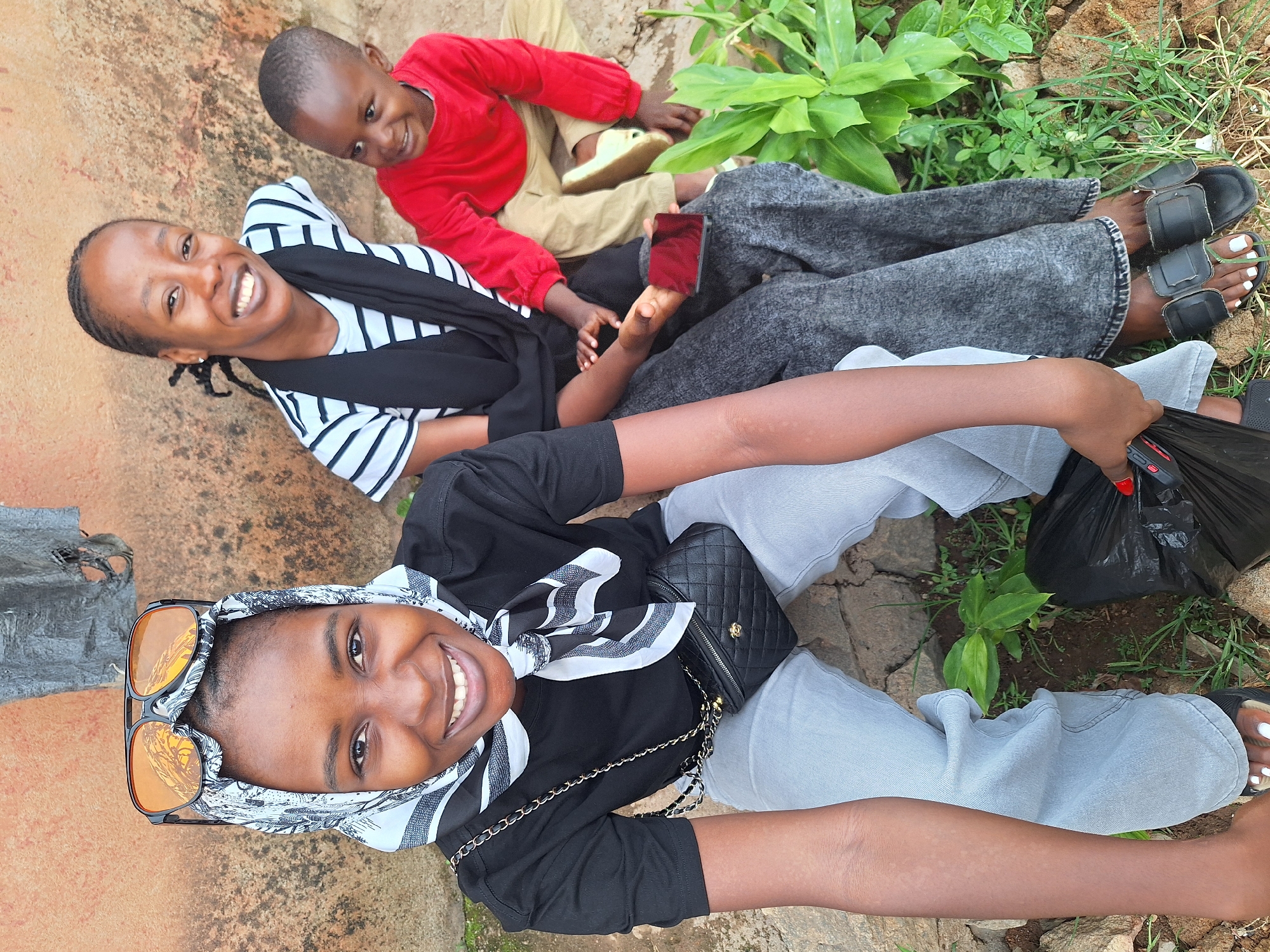 The image depicts three individuals outdoors in Sabon Tasha, Nigeria, positioned against a textured wall and natural ground. In the foreground, a woman with dark complexion smiles towards the camera, wearing a black short-sleeved top, light blue jeans, and a white and black patterned headscarf. Orange-lensed sunglasses rest on her headscarf. She wears a black quilted crossbody bag and holds a black plastic bag in her left hand, which has red nail polish; her visible right foot displays white nail polish. To her right, a second woman with a dark complexion and braided hair also smiles at the camera, dressed in a white and black striped top, a black shawl, and dark grey acid-wash style pants. She holds a dark-colored smartphone with a red reflection on its screen; her feet are in black slide sandals with white nail polish. To the right of the second woman, a young boy with a dark complexion, wearing a red long-sleeved shirt and light brown pants, smiles. The background features an orange-brown and grey textured wall, with a dark, tattered object visible in the upper center. The ground is an uneven mixture of earth and rocks, interspersed with green plants and grass.