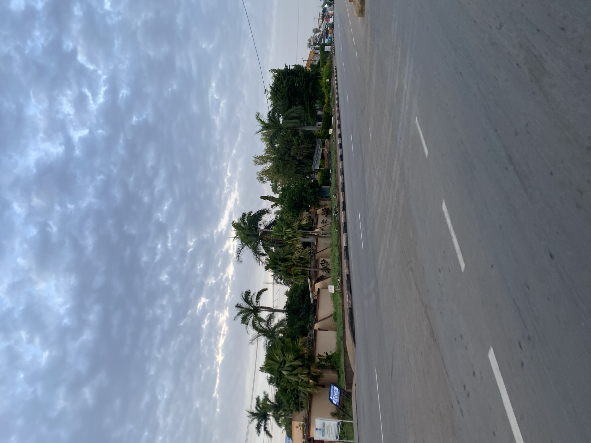 An exterior, eye-level, medium shot shows a street scene in Jos, Nigeria, under a cloudy sky. The street in the foreground is paved and has white lane markings. To the left of the street is a beige-colored wall topped with barbed wire. Behind the wall, several palm trees and dense green foliage are visible. Signs are posted near the wall, one of which appears to be a white informational sign. Further down the street, buildings and more trees can be seen. The sky is filled with dramatic, textured clouds. The scene is static and depicts an urban environment.