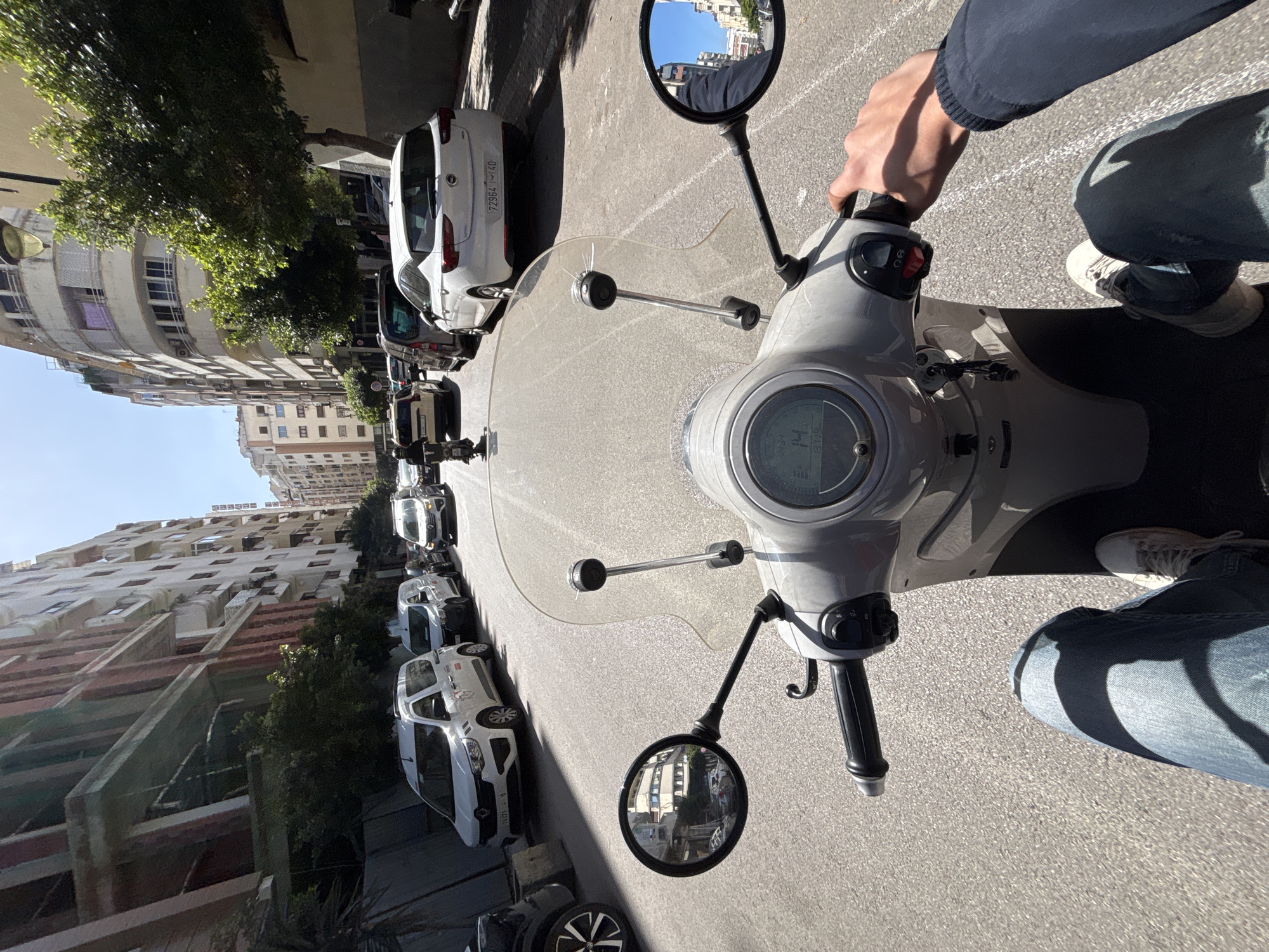 The image provides a first-person perspective from a light-grey scooter on an asphalt street in Tanger, Morocco, under bright daylight with a clear blue sky. The scooter's transparent windshield, both rearview mirrors, and a digital dashboard displaying "14" (indicating speed) and "13.78" are visible. The rider's legs, clad in blue jeans and white sneakers, and their right hand gripping the handlebar are prominent in the foreground. A set of keys hangs from the ignition.
The street ahead is occupied by vehicular traffic. Directly in front of the scooter is a white Opel Astra hatchback with Moroccan license plate "72964 | 40". Behind it, a queue of additional cars and a smaller scooter with a helmeted rider are visible. The left side of the street features multiple parked white vans, including one with a partial Moroccan license plate "747...8", positioned alongside a multi-story building under construction with exposed concrete and red vertical architectural elements, partially obscured by green bushes at ground level. Beyond this, older multi-story residential buildings with balconies are visible. The right side of the street is lined with multi-story residential buildings characterized by cream-colored facades and curved balconies, interspersed with trees and a tall street lamp extending over the roadway.