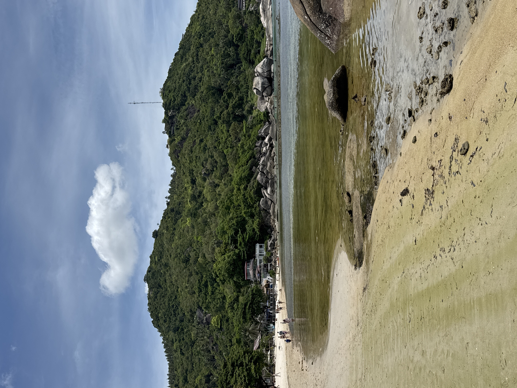 The image depicts a beach scene in Baan Chaweng Noi, Thailand. A densely vegetated hillside slopes down to a sandy beach and shoreline. A communications tower is visible on the upper part of the hill. Several structures are situated along the base of the hill. People are scattered along the shoreline and interacting in small groups. The water is murky in the foreground. Large rocks are present both in the water and on the beach. The sky is blue with a few scattered clouds.
