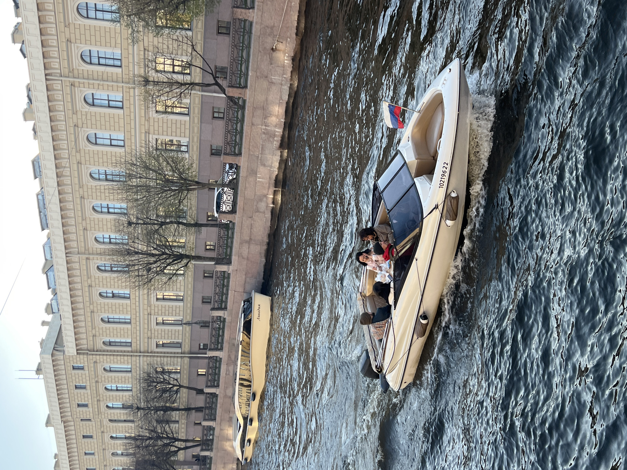 The image captures a scene in Saint Petersburg, Russia, likely along one of the city's canals. A motorboat is moving through the water, carrying several passengers. A Russian flag is displayed on the boat. The side of the boat has the number "10219622" printed on it. Adjacent to the canal is a multi-story building with numerous arched windows and a light-colored facade. Bare trees stand along the canal bank in front of the building. A second, similar boat is moored along the bank to the left of the moving one, bearing a word that appears to read "Лахадия" (Lakhadiya in transliteration). The water has a dark, reflective quality.