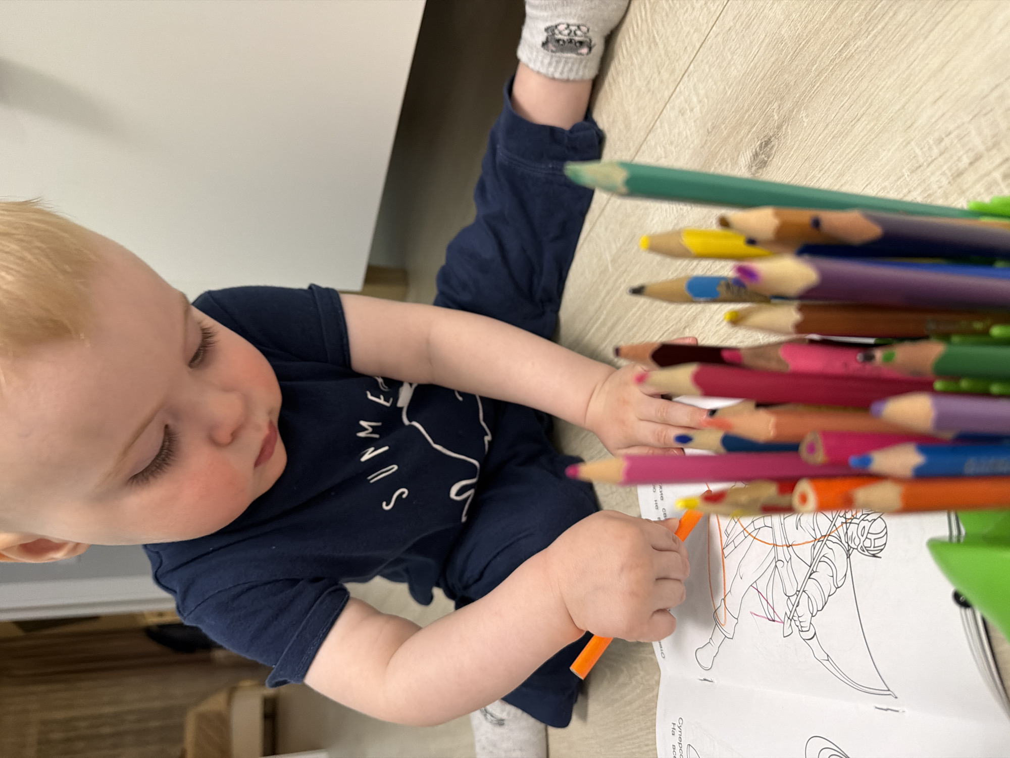 An overhead view captures a young, fair-skinned child with blonde hair intently coloring in a book. The child, likely a toddler, wears a dark blue t-shirt, partially revealing the word "SUMME" (presumably "SUMMER"), and grey socks featuring small dog faces. An orange colored pencil is held in the child's left hand, actively filling in an outline drawing of a warrior-like figure, possibly wielding a scythe or staff, within an open coloring book.
To the child's right, a substantial collection of brightly colored pencils, including green, yellow, blue, purple, brown, pink, red, and orange, is fanned out, with some housed in a green plastic holder. The coloring book page displays various outline elements and partial Russian text, specifically "Суперсо," "На," "все," and "отло," alongside the numeral "1."
The scene is set indoors, with a light wooden surface serving as a ground or low table, and a plain white wall or furniture piece forming the background. The bright, even lighting suggests a well-lit interior, potentially in Votkinsk, Russia. The child's focused expression and calm demeanor indicate deep engagement in the creative activity.