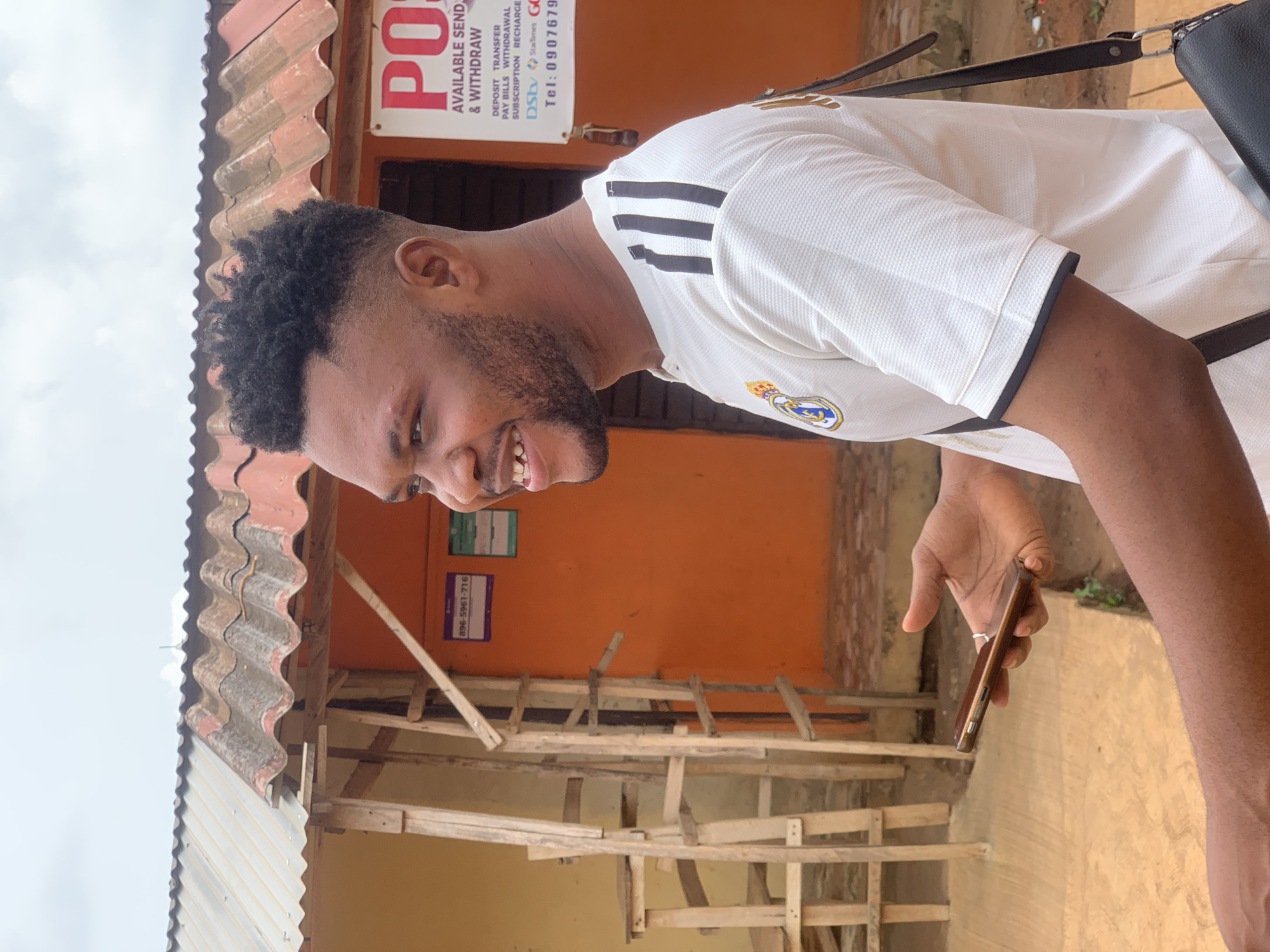 A young man with short, dark, curly hair and a slight beard is smiling and looking down at a brown smartphone in his right hand. He is wearing a white Real Madrid soccer jersey with black stripes on the shoulders and a black collar. A black bag is slung over his shoulder. He stands in front of a building with an orange facade and a corrugated metal roof. A wooden structure, possibly scaffolding, is in the foreground. A sign with the letters "PO" is visible, advertising money transfer services. A phone number is on the sign, suggesting the location is Ikorodu, Nigeria.