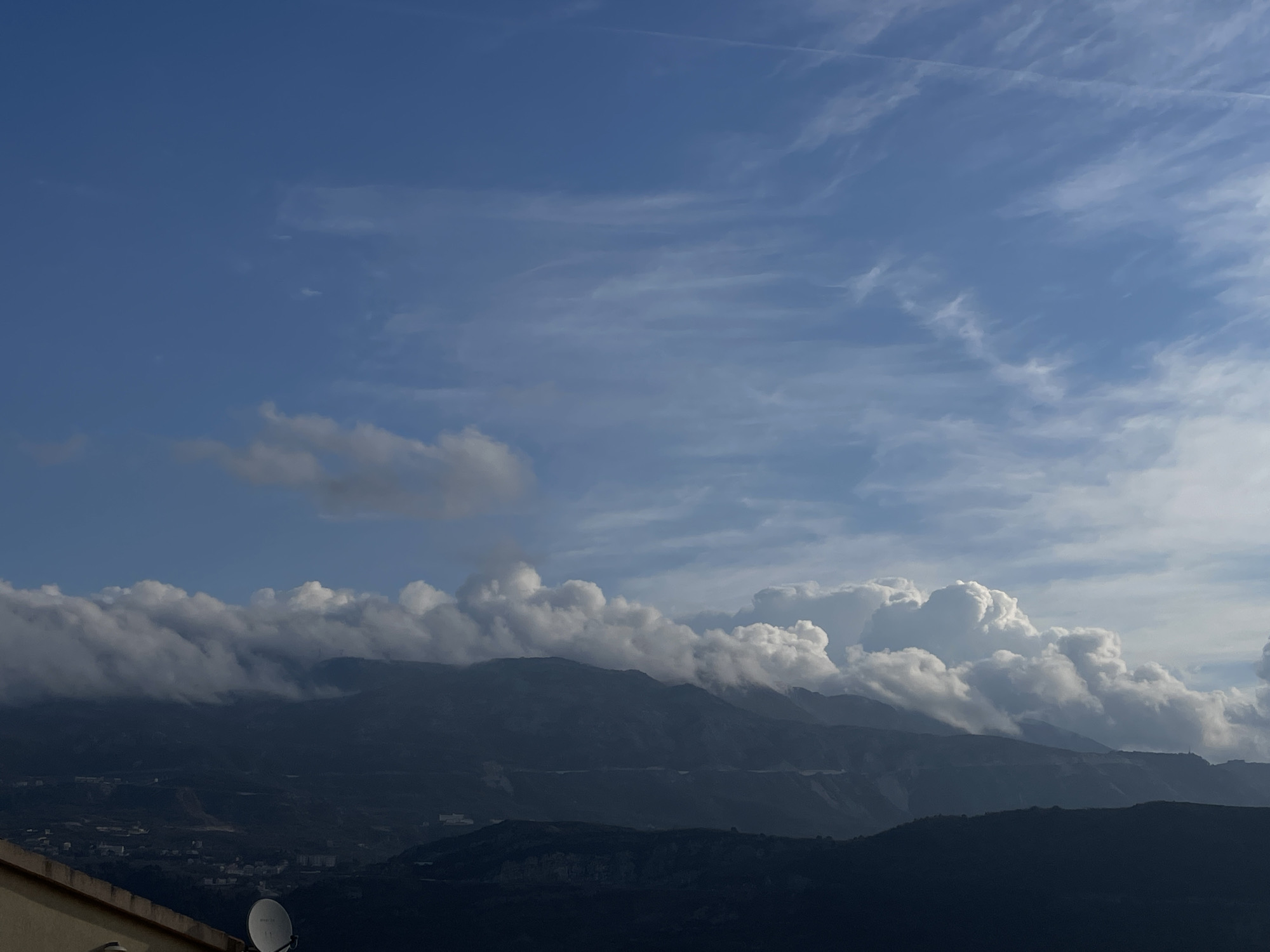 The image presents a mountainous landscape consistent with Budva, Montenegro, under a partly cloudy blue sky. The upper sky displays clear blue expanses with scattered, wispy white cirrus clouds and a single faint contrail. Below this, a significant band of white and grey cumulus clouds is visible, resting on and partially obscuring the dark, rugged mountain ranges that extend across the middle ground and background. The mountains appear layered, with visible slopes and peaks. In the lower-left foreground, a section of a light-colored building with a defined roof edge is present, upon which a white, circular satellite dish is mounted, angled upwards. Below the foreground structure and receding into the mid-ground, a populated area characterized by numerous small, indistinct buildings is discernible on the lower mountain slopes. No individuals or specific actions are depicted. The scene is illuminated by natural daylight.