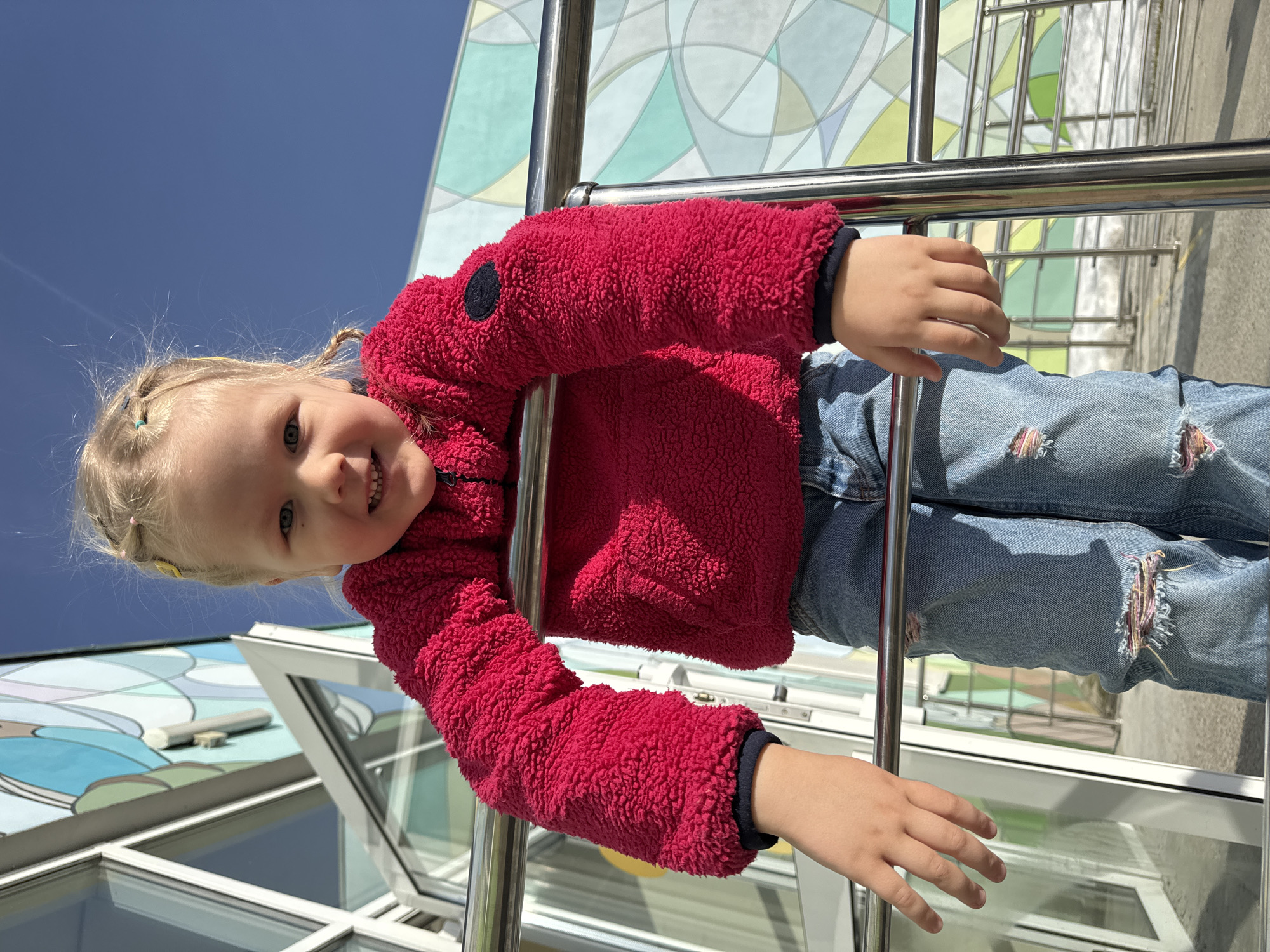 A young, fair-skinned girl with blonde hair braided into pigtails stands beside a railing. She wears a red fleece jacket and blue jeans with torn knees. The background features a building with a colorful facade under a bright blue sky. The location is Perm, Russia.
