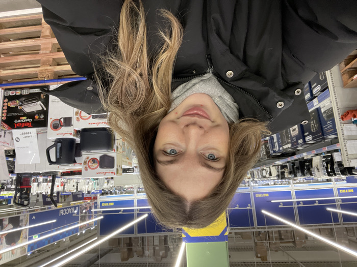 The image shows a person taking a selfie in what appears to be a large retail store, possibly a hypermarket or a warehouse club, as suggested by the visible "METRO" signage. The image is oriented upside down, with the person's head at the bottom.
The central subject is a young person, likely female, with fair skin, light-colored eyes (blue or grey), and long, wavy or slightly curly light brown hair with blonde highlights, which falls around her face and shoulders. She is wearing a grey hooded sweatshirt under a black puffer jacket with a high collar. Her expression is neutral, with a slight hint of a smile. Her lips are a natural pink color.
The background reveals rows of store shelves stocked with various products, predominantly kitchen appliances. To the person's left, an aisle features boxed items, including a "Tefal" brand toaster or grill with a price tag showing "14990" (possibly Russian Rubles, with a strikethrough price of "22990"). Below this, there are smaller black appliances, possibly kettles or coffee makers, from the "Moulinex" brand, packaged in white and red boxes. A black mug with a handle is visible in front of one of these boxes.
To the person's right, more shelves are filled with boxed goods, some appearing to be coffee makers or other small kitchen electronics from brands like "De'Longhi." The shelving units are metal, typical of a retail environment.
Further in the background, parallel to the aisles, there's a blue structure with white text that clearly reads "METRO" several times, indicating the name of the store. Below these blue signs, there are long, linear fluorescent lights running across the ceiling, illuminating the store. On the left side of the image, another blue sign with white text is visible, which appears to read "КАССЫ" (KASSY), meaning "Checkouts" or "Cashiers" in Russian. The floor is light-colored.
On the far left, partially obscured, is a stack of wooden pallets, suggesting goods are stored or displayed in a warehouse-style setting. The overall impression is a casual, everyday shopping trip in a large, well-lit retail space.