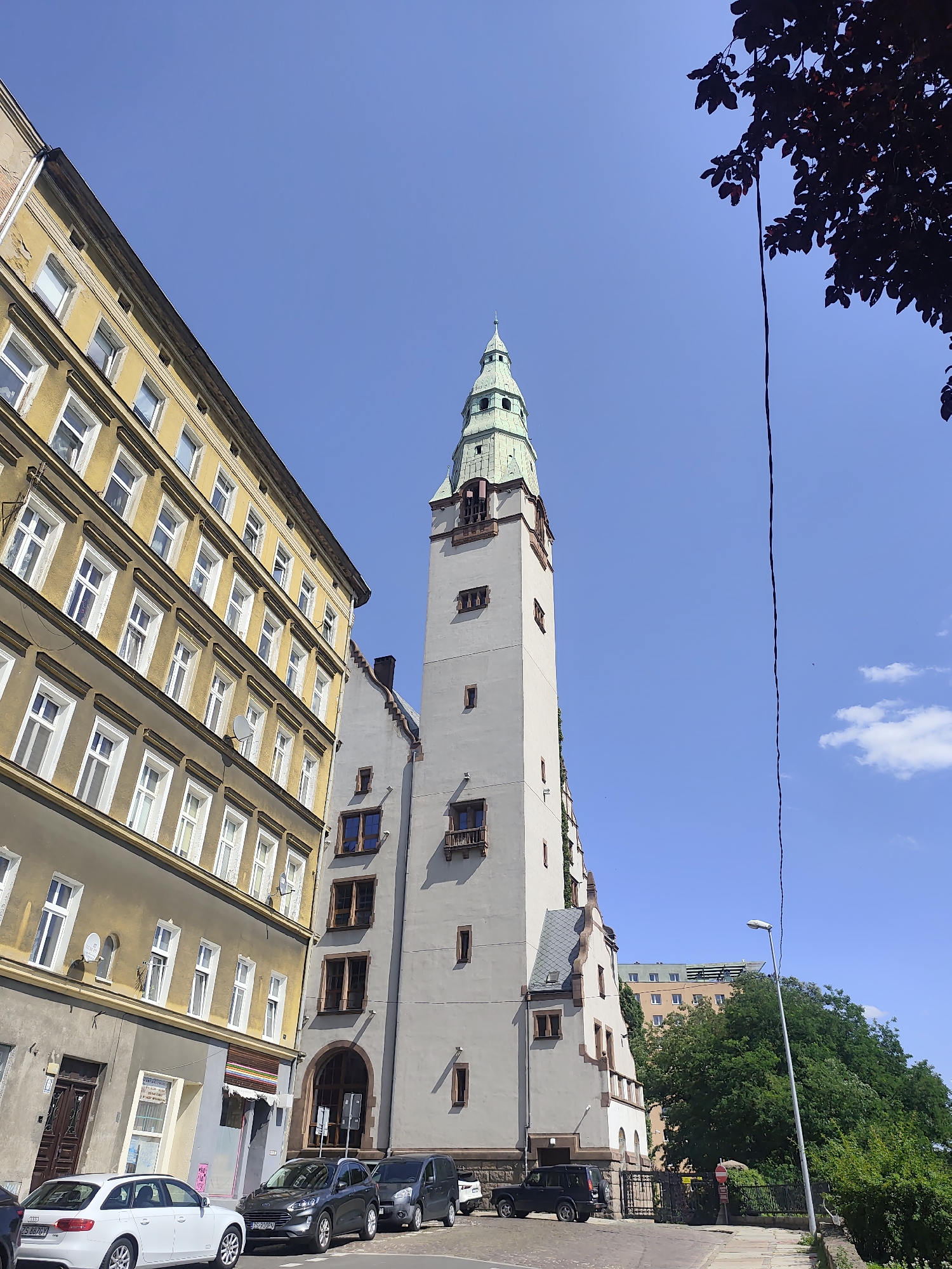 The image captures a street scene in Szczecin, Poland, featuring a prominent tall, light-colored building with a green-capped spire, characteristic of the Old Town architecture. To the left, a yellow building with multiple stories and uniformly spaced windows displays signs of age. In the foreground, several parked cars are visible on a cobblestone street. The sky is a clear blue, with a few wispy clouds. The scene suggests a typical urban environment on a sunny day.