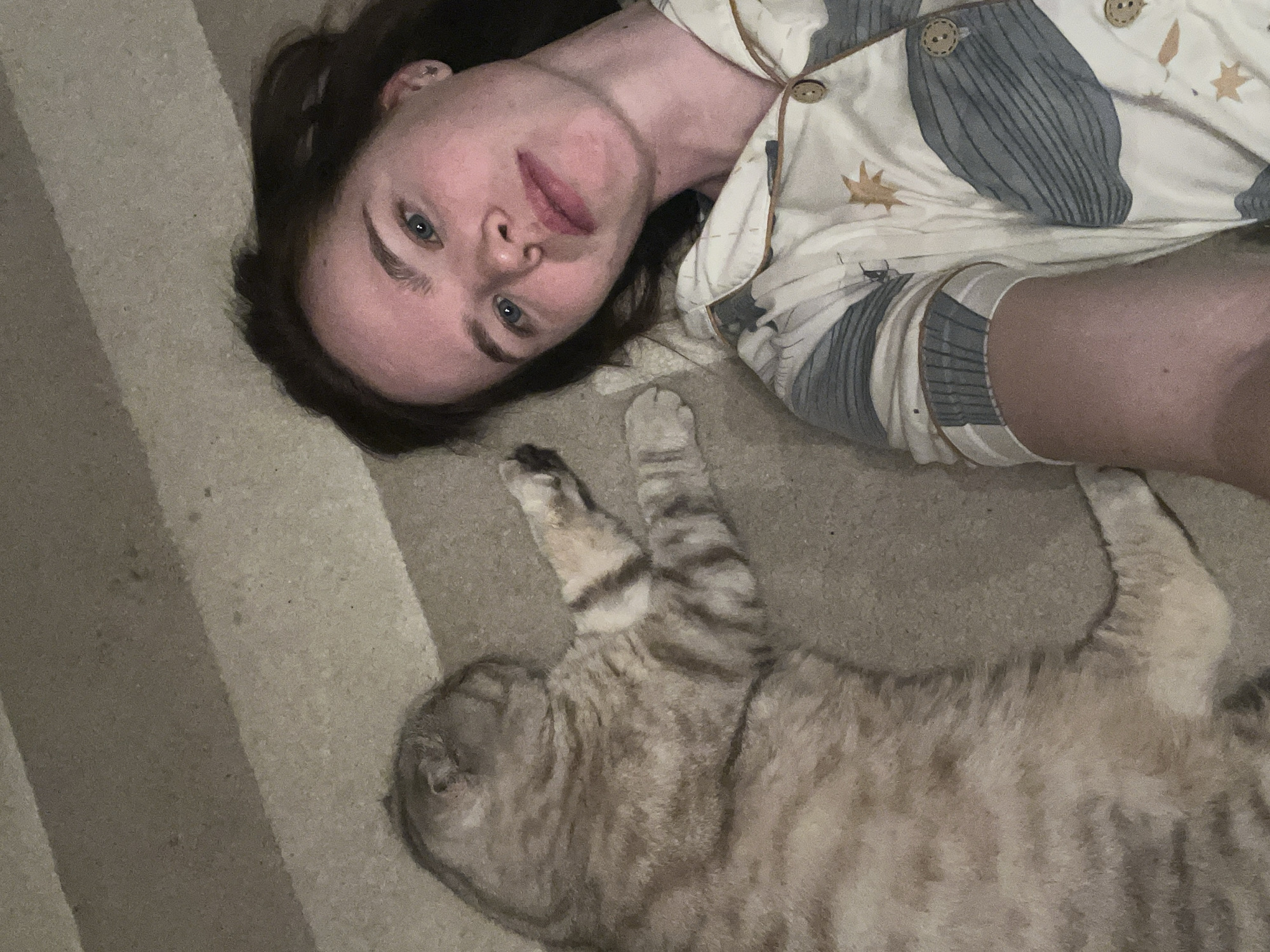 A woman is lying on a striped carpet next to a tabby cat. The woman is wearing pajamas with a pattern of whales and stars. The cat is lying on its side with its paws outstretched. The location is unknown.