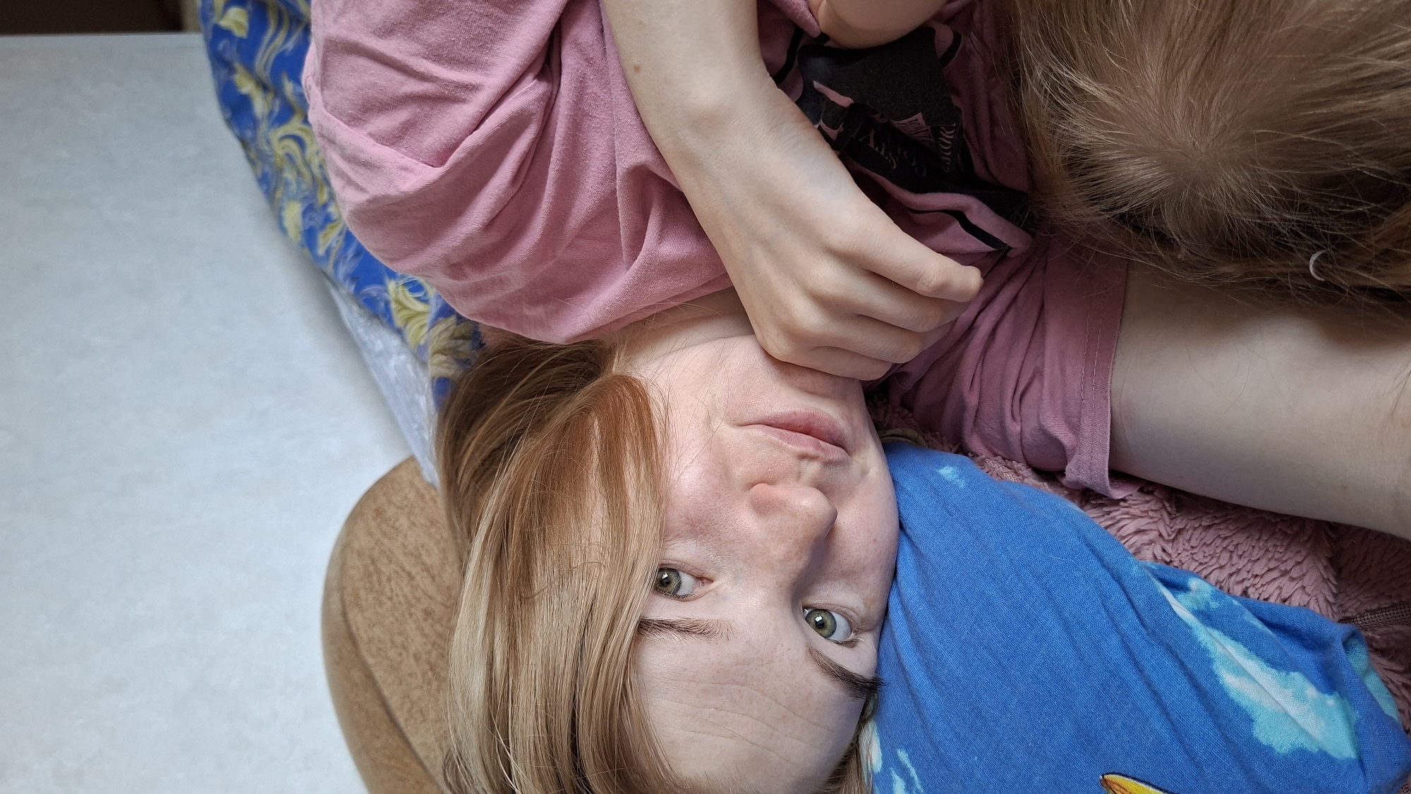 A woman with light skin and blonde hair is lying on her back, looking into the camera. She is wearing a pink shirt and has a blue cloth near her face. A child with light brown hair is lying next to her, possibly on a pink blanket. The woman is holding the child with her hand. They are positioned on a surface covered with a blue and gold patterned cloth. The location is not discernible from the image.
