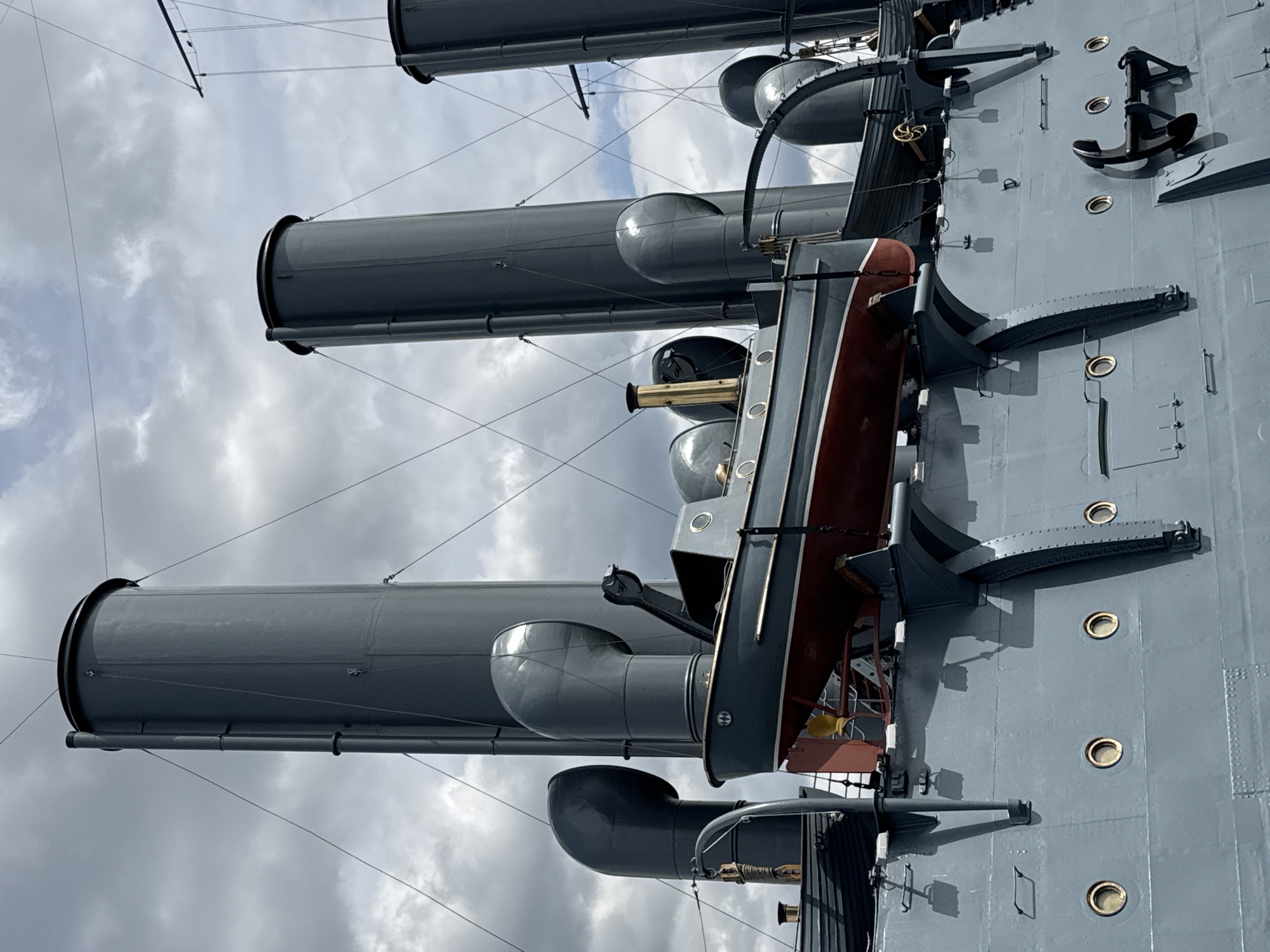 This close-up image depicts a significant portion of a large, grey warship's upper deck and superstructure, likely a historical vessel. The main subjects are three prominent, cylindrical grey smokestacks that rise vertically towards a cloudy sky. Their tops feature a dark, possibly black, rim.
Between the smokestacks and the ship's side, a smaller boat, painted dark grey with a reddish-brown lower hull, is suspended from davits. This appears to be a ship's launch or a large lifeboat, indicating the ship's operational capabilities or historical configuration. Below the suspended boat and along the ship's grey hull, several circular portholes with distinct bright, possibly brass or gold-colored, rims are visible. Further forward on the deck, a large black anchor is prominently displayed and secured.
Numerous other structural details are present, including various grey vents, pipes, and support structures around the smokestacks and on the deck itself. Thin wires or rigging lines stretch across the frame, suggesting masts or antenna arrays.
The scene is set during the daytime under an overcast sky, characterized by soft grey and white clouds. There is no human activity visible, suggesting the ship is stationary, possibly docked or moored as a museum ship. Given the location context of Saint Petersburg, Russia, this strongly implies a historical naval vessel. The ship's design and color scheme convey a sense of strength and historical industrial marine engineering. There is no visible text.
