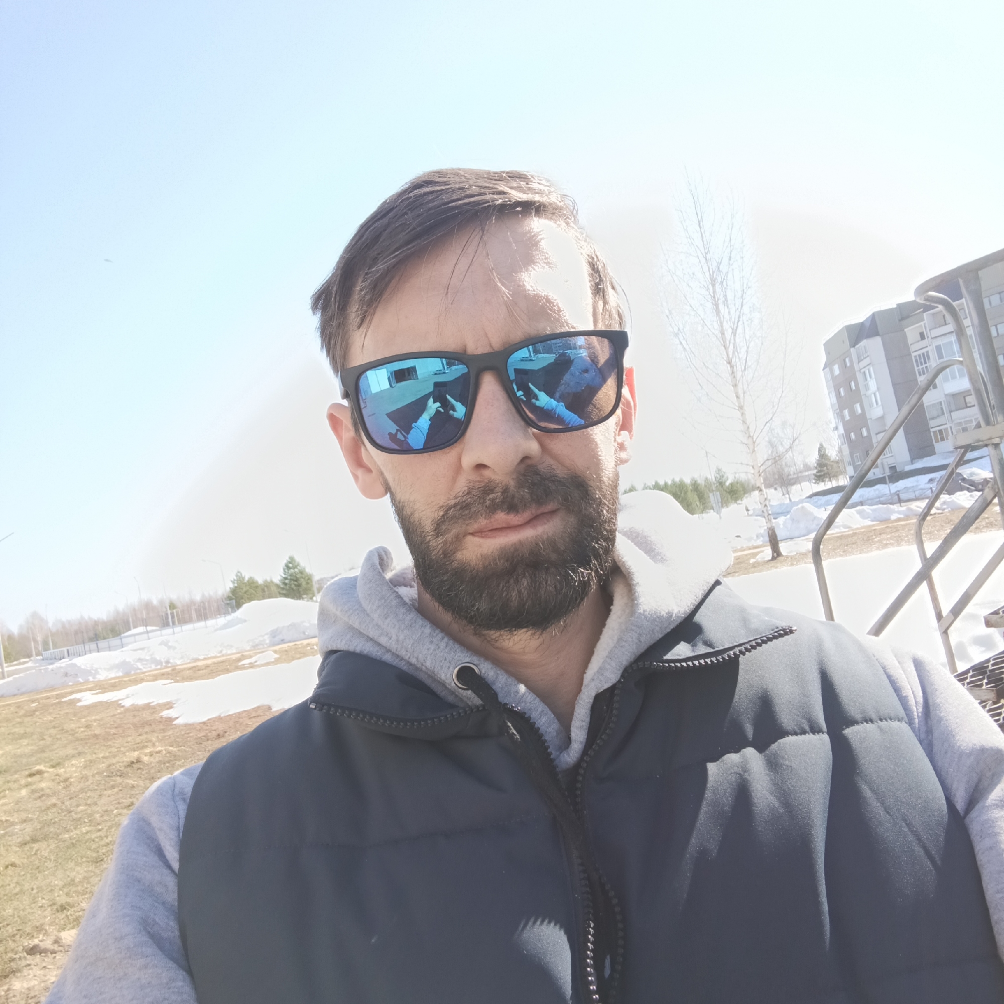 A man with a beard and blue mirrored sunglasses is taking a selfie outdoors. He's wearing a gray hooded sweatshirt and a navy blue vest. The reflection in his sunglasses shows a hand holding a phone. The background includes melting snow, a grassy area, trees, a building, and some metal stairs. The photo was taken in Марковский, Russia.