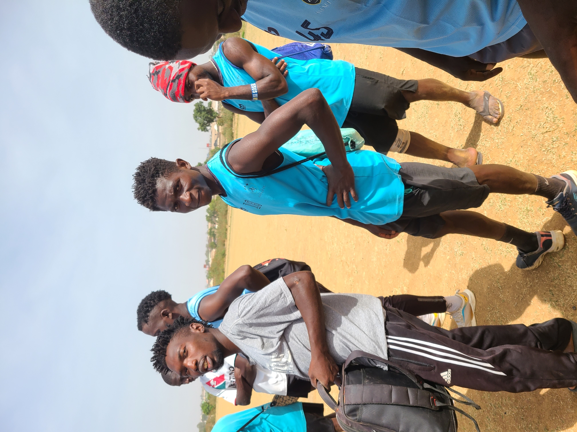 The image shows a group of young men standing outdoors, likely in Barikin Jirga Summa, Nigeria, based on the light and background vegetation. The men are dressed in a mix of athletic wear, including light blue jerseys, shorts, sweatpants, and t-shirts. One man wears a red and white head covering. Some are wearing athletic shoes and another is wearing sandals. One man is carrying a backpack. They appear to be a team or group of friends, possibly after a sports activity. The setting is a dusty, open area with low-lying buildings and vegetation visible in the background under a clear sky.