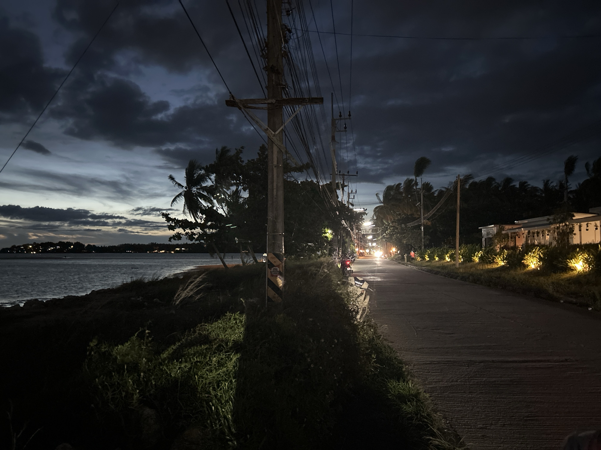 The image shows a nighttime scene in ตำบลเกาะพะงัน, Thailand. A coastal road runs alongside a dark shoreline. On the left, the ocean is visible with lights on the distant land. Tall grass and vegetation grow along the bank. Power poles and lines stretch across the road overhead. On the right, a road is lined with landscaped plants and building with lit exterior. The road appears to be wet. Car and motorcycle headlights are seen in the distance along the road. The sky is dark with clouds.