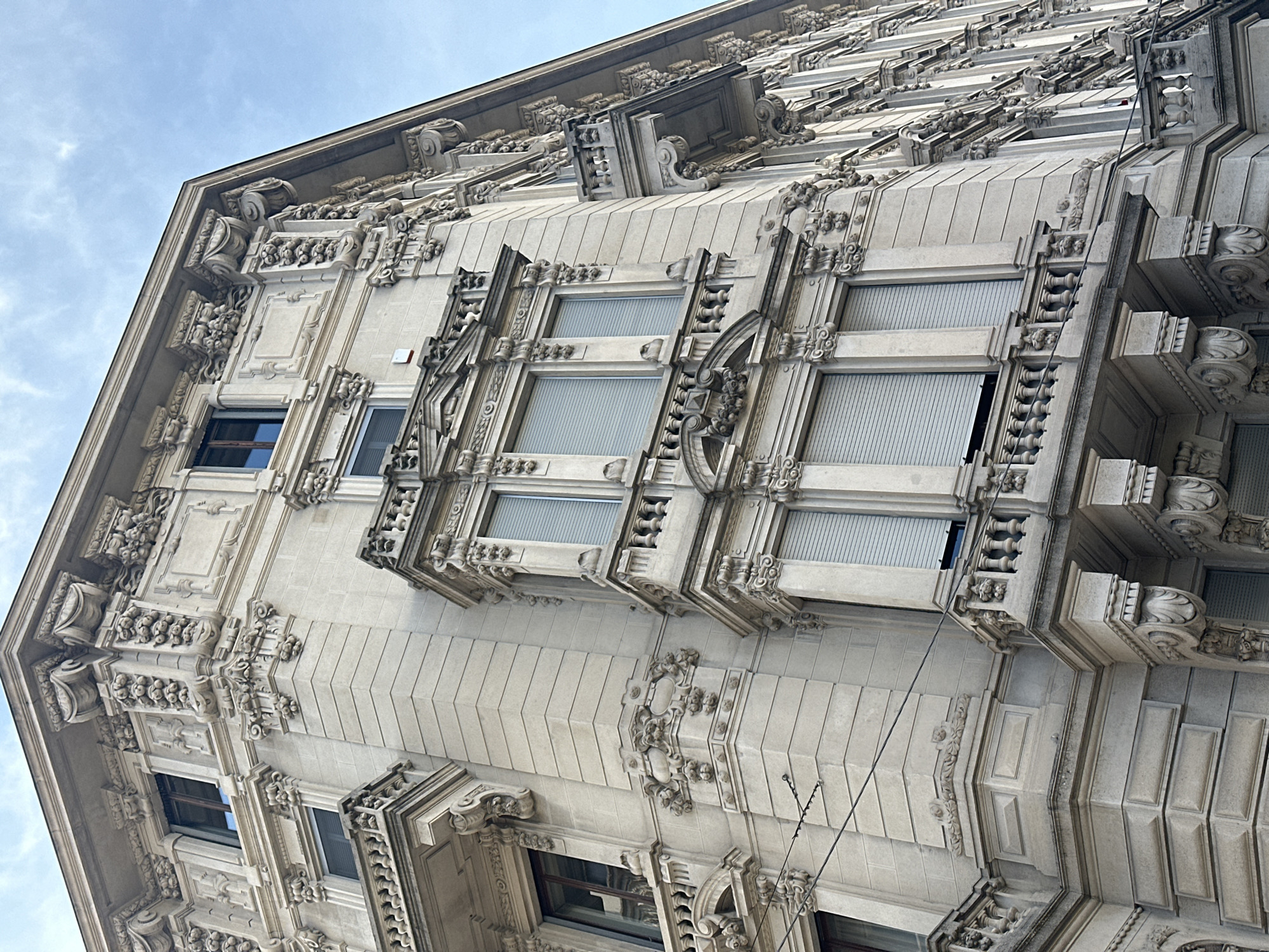 An ornate, multi-story building facade in Milan, Italy, is captured from a low-angle perspective, emphasizing its corner and upper levels against a bright, partly cloudy blue sky. The edifice is constructed of light-colored stone, extensively adorned with classical and Art Nouveau-inspired sculptural details including elaborate balustrades, pilasters, cornices, floral garlands, and volutes. Prominent bay windows characterize the corner, flanked by standard rectangular windows, some equipped with light-grey roll-down shutters, while others display dark-framed panes, with a few partially open. Small, individual balconies with matching balustrades project from the side facade. Overhead utility wires traverse the upper left of the frame. No individuals, vehicles, or specific street features are discernible.