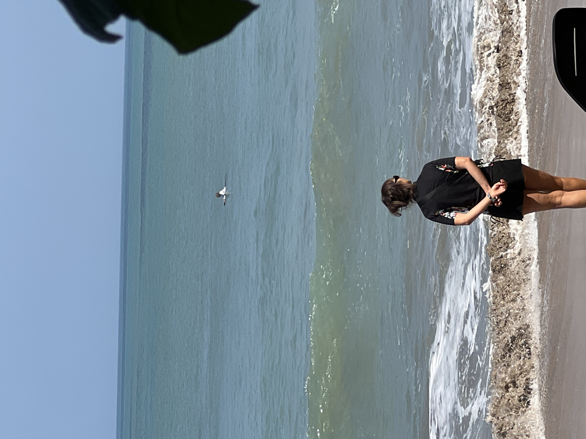 A woman stands on a sandy beach in Kuta, Indonesia, facing the ocean. She is wearing a black rash guard with floral accents and a black skirt. Her hands are clasped behind her back, and she is wearing sunglasses. The ocean is a calm, teal color with small waves breaking near the shore. In the distance, a person on a surfboard can be seen in the water. A dark green leaf is visible in the upper right corner of the image, partially obscuring the view. The sky is a clear, pale blue.