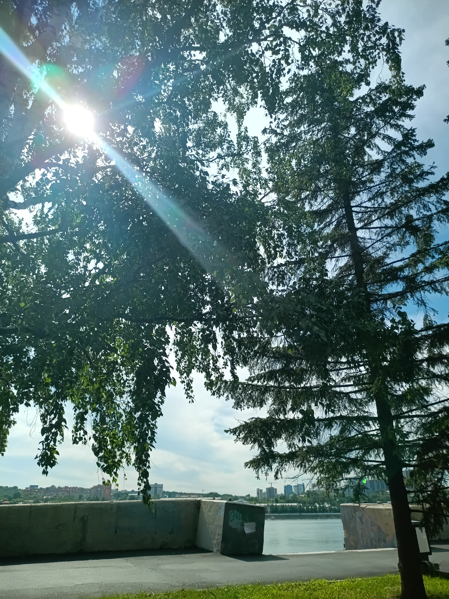 This image depicts a sun-drenched scene with dense green foliage from trees in the foreground, partially obscuring a bright sky with scattered clouds. Sunlight beams through the leaves, creating lens flare effects. In the middle ground, a concrete barrier runs horizontally, with a body of water visible beyond it. Further in the distance, the cityscape of Irkutsk, Russia, is visible, featuring a skyline of buildings and trees lining the opposite bank of the river. The overall impression is one of a bright, sunny day in an urban environment with natural elements.