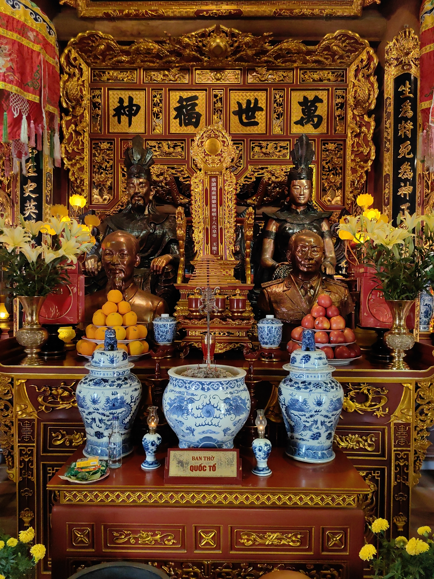 The image shows a religious altar in a building located in Hong Gai Ward, Vietnam. The altar is heavily ornamented with gold trim and includes statues of figures with ornate headdresses. Busts of other figures stand in front of the statues. There are offerings of fruit, vases of flowers, and ceramic urns decorated in blue and white. A red and gold sign bearing Vietnamese script reads "BAN THO QUỐC TỔ."