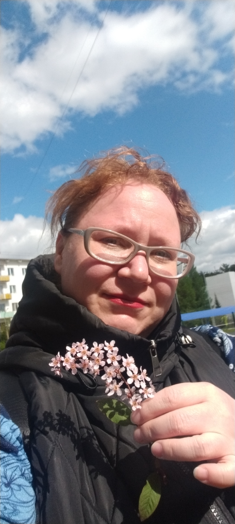 A woman with reddish-brown hair, wearing glasses and lipstick, is holding a sprig of small pink flowers in her right hand. She is wearing a black jacket. The background shows a bright blue sky with white clouds. Part of a light-colored building is visible to the left of the woman's head. The location is Chaikovsky, Russia.