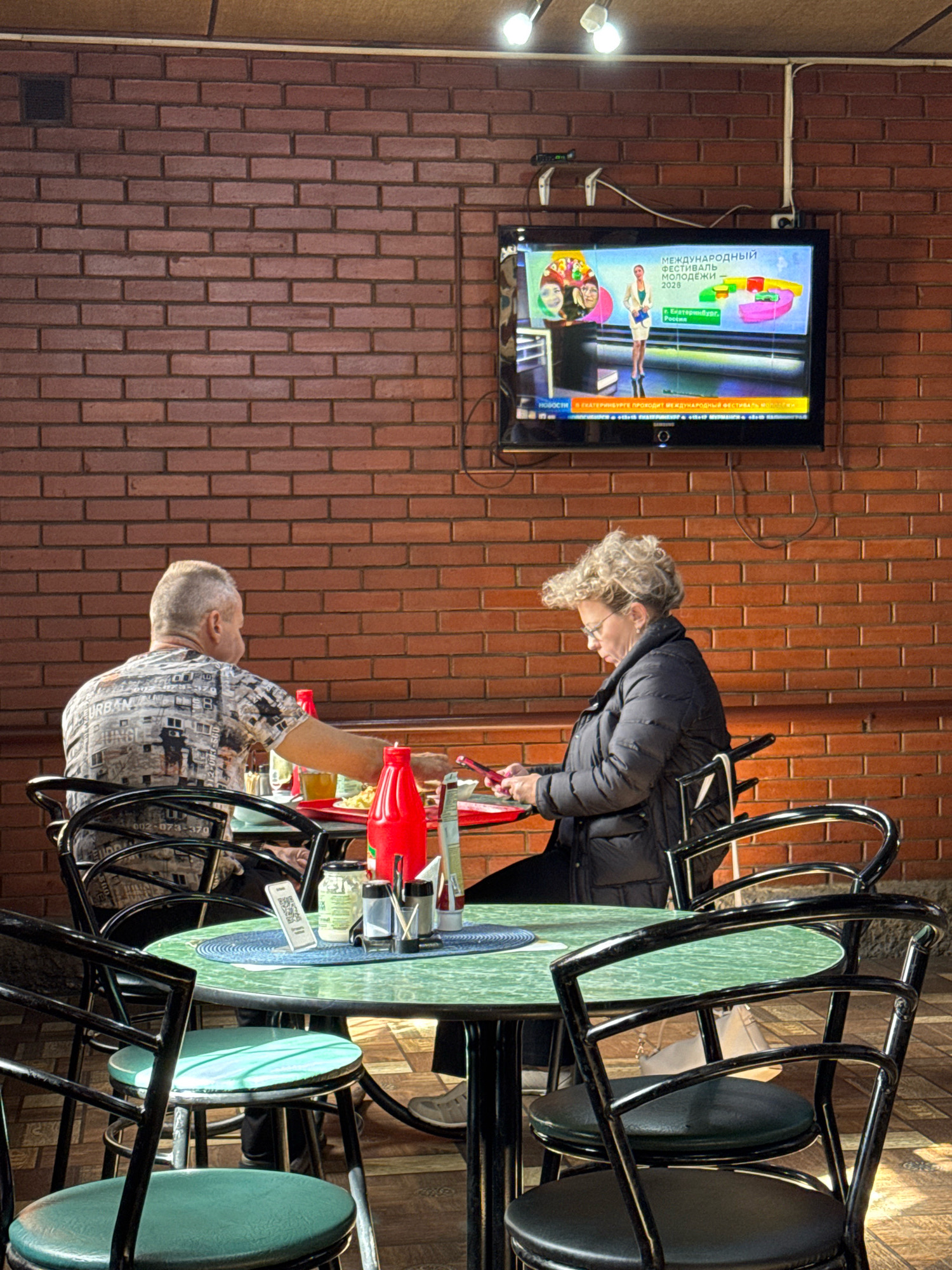 This image depicts an outdoor cafe setting with a brick wall as the backdrop. Two individuals are seated at a table. On the left, a man with gray hair is looking towards the table, and on the right, a woman with blonde curly hair and glasses is looking at her phone. The table is set with plates of food, drinks, condiments, and a QR code stand. A television screen mounted on the brick wall is displaying a news broadcast. The overall environment suggests a casual dining experience. The image does not contain information that allows for localization to Novyi Berezai, Russia.