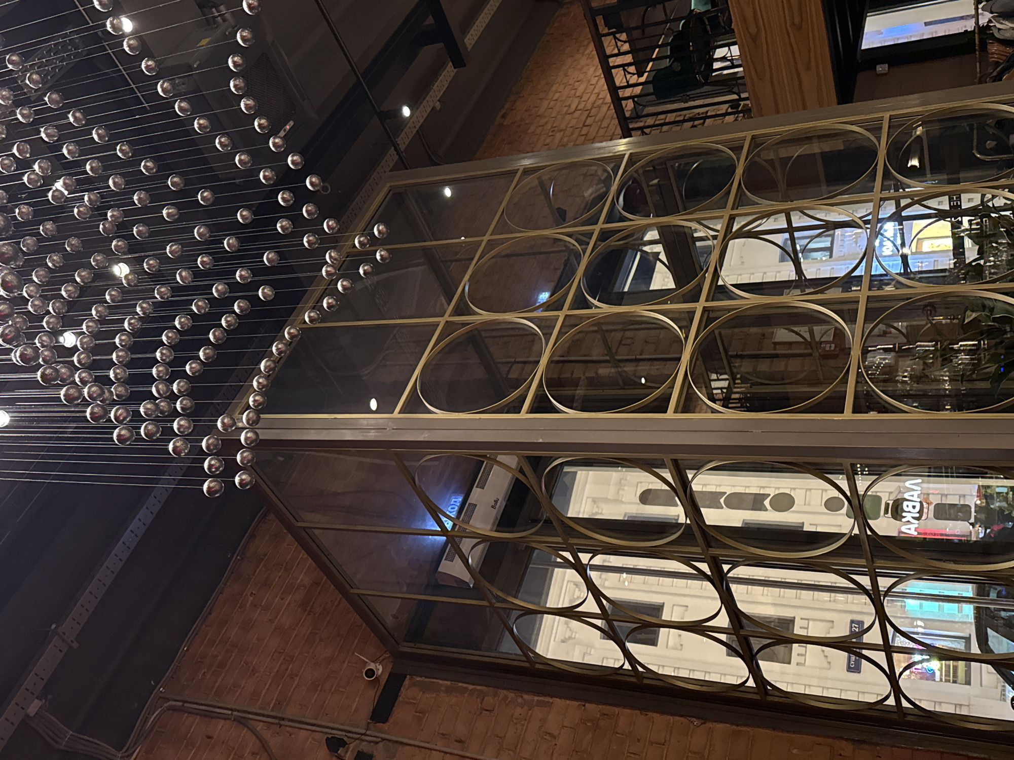 The image shows an interior view, looking up at a decorative feature composed of a grid of glass panels framed by gold-colored metal circles and straight lines. To the left is a large lighting fixture made of many small, spherical bulbs suspended on thin wires. The background reveals exterior buildings, including a sign written in Russian that says "АКВАЛ" (AKVAL), and a street sign that partially reads "СУШ... 27" (SUSH... 27), suggesting the presence of a sushi restaurant. The brick wall visible at the bottom suggests that this location is in Moscow, Russia. A security camera is also visible on the brick wall.