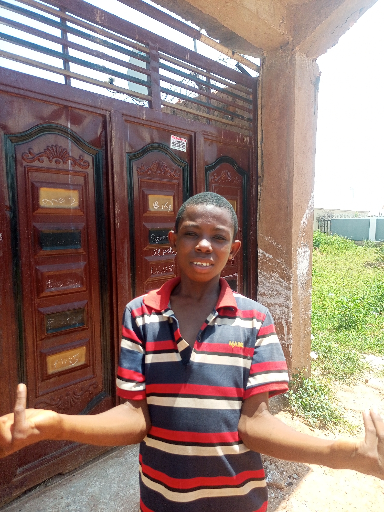 A young boy stands in front of a brown, ornate double door, his arms outstretched. He is wearing a horizontally striped polo shirt with red, white, and blue bands, and the word "MAKS" is visible on the left chest. His left hand is raised with two fingers extended, while his right hand is out of frame. The doors have decorative carvings and several white markings, possibly graffiti. To the left of the door, the boy is standing next to a rough, textured, light brown concrete pillar. Behind the pillar, a grassy area with some weeds and a distant fence is visible under a bright sky. The scene appears to be outdoors in a residential or public area.