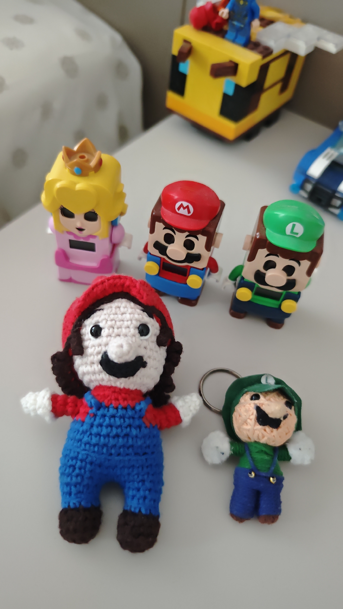 The image shows a white table surface upon which are arranged several Super Mario Bros. themed objects. In the background, there are Lego versions of Princess Peach, Mario, and Luigi. In the foreground are crocheted versions of Mario and Luigi. The crocheted Luigi has a keychain ring attached. The background also shows a Lego model of a Minecraft bee with a blue minifigure on top of it and the front of a blue toy car. The location of the scene is not discernable in the image. The location of the city of Burgas, Bulgaria is also not discernable in the image.