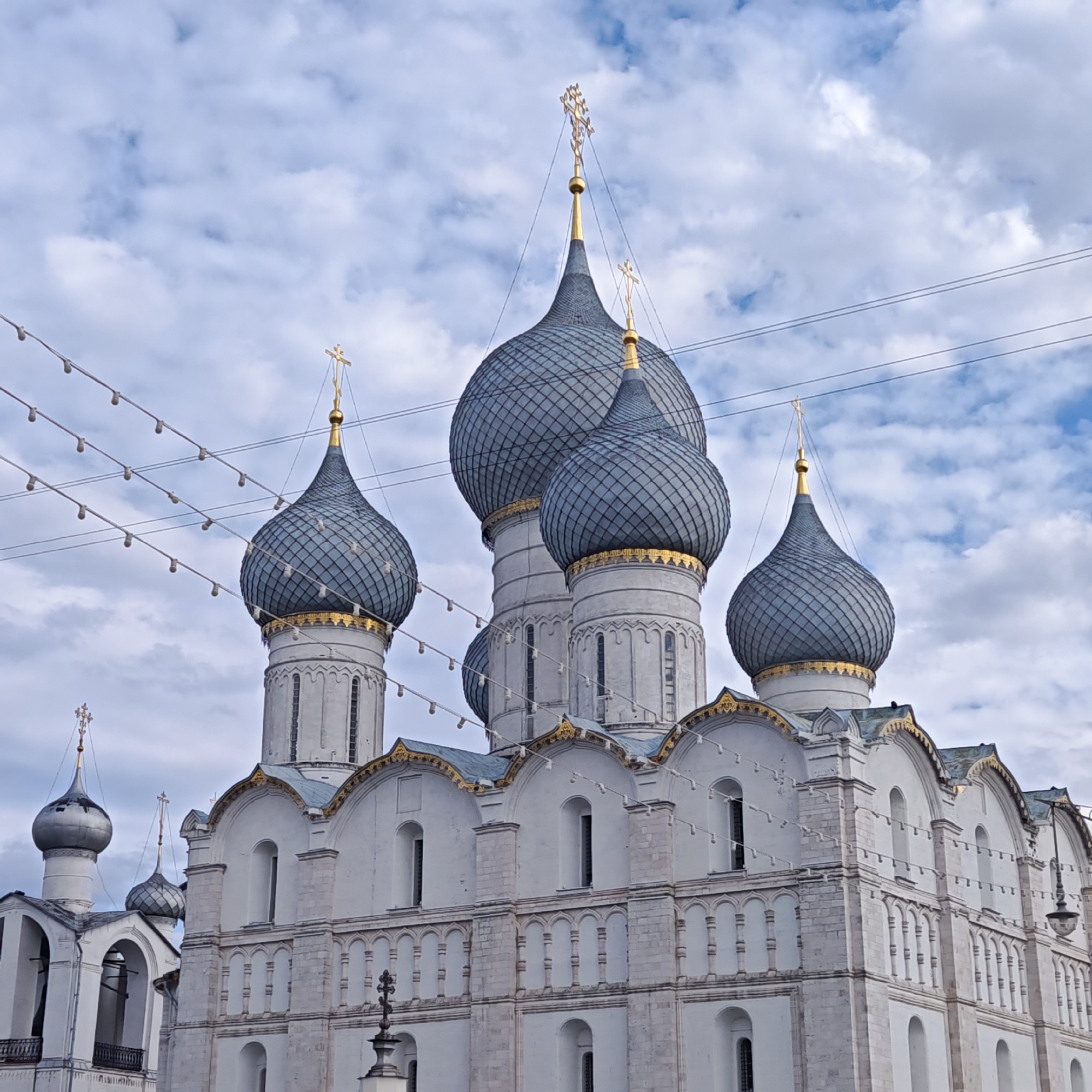 The image depicts a Russian Orthodox church, possibly located in Rostov, Russia, given the architecture and general style. The building is white with intricate architectural details, including arched windows, decorative trim, and multiple levels. It has several onion-shaped domes painted in gray with a diamond pattern. Each dome is topped with a golden cross. The sky in the background is light blue with clouds. Festoon lighting (strings of lights) is strung across the foreground of the image.