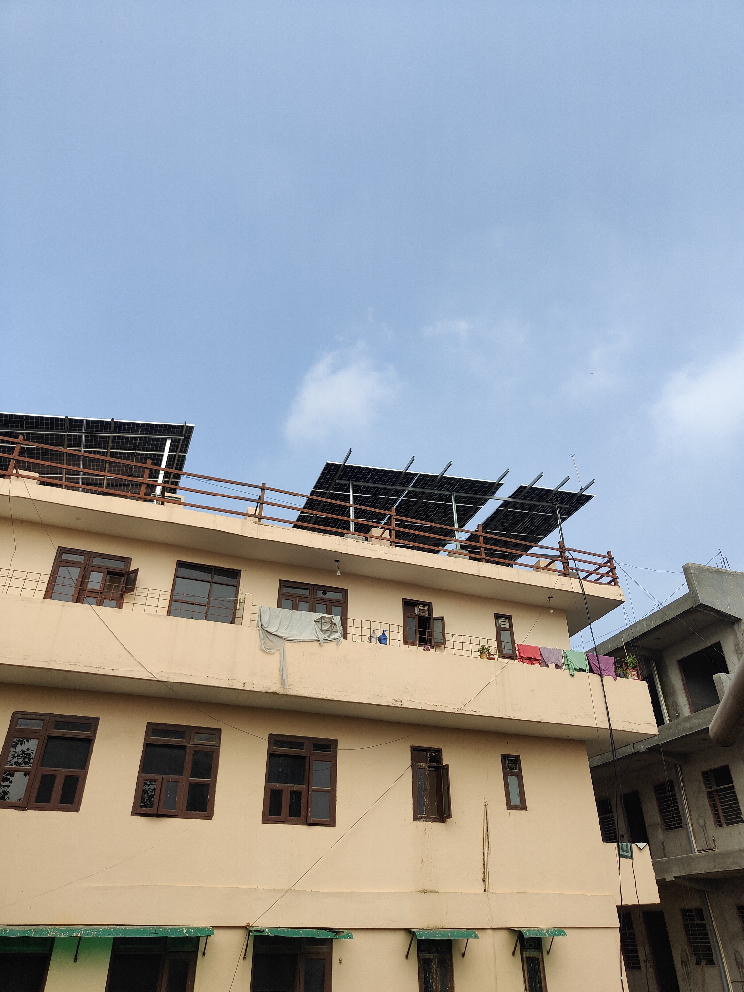 This is a medium-angle, low-rise exterior shot of a beige apartment building, situated in the city of Gurgaon, India. The building features multiple levels of windows with dark brown wooden frames. Some windows are open, revealing glimpses of the interior. On the roof, a set of solar panels are installed on a metal frame, with a wooden railing encircling the rooftop. Clothes are also seen hanging to dry on a clothesline on the rooftop balcony. Adjacent to the beige building is a partially constructed grey concrete building. The sky is a clear blue with sparse white clouds.