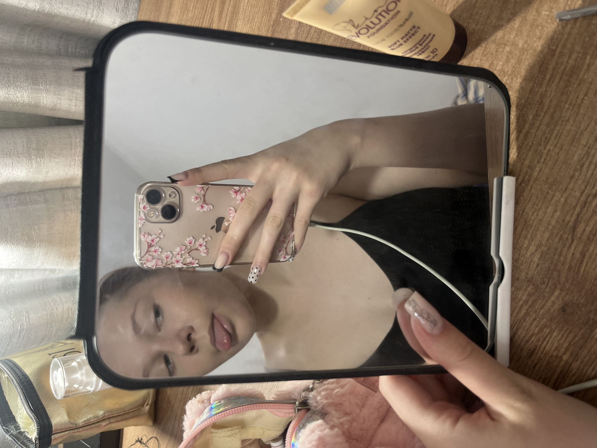 A young person, likely female, is captured in a mirror selfie, holding a smartphone with a clear case adorned with a pink floral design. Their face is visible in the reflection, looking sideways with their tongue playfully sticking out. They are wearing a black top.
The mirror, with a black frame, is positioned on a light-colored wooden surface, which appears to be a desk or table. In the upper right corner, outside the mirror's reflection, lies a yellowish tube of cosmetic product with the visible text "EVOLUTION," "FOUNDATION," "SOFT MATTE BLUR EFFECT," and "КРЕМ ТОНАЛЬНЫЙ" in Cyrillic. Other items visible on the wooden surface outside the mirror include a gold makeup bag with a zipper, a small clear bottle, and a pink fluffy pouch with an iridescent trim. Inside the mirror's reflection, a light-colored, textured curtain or fabric is visible in the background.
The person's hands are prominently featured, showing long, elaborately manicured nails. Some nails have black tips, one has white polka dots on a black background, and others feature intricate designs. The Apple logo is partially visible on the phone case. The scene depicts an indoor setting, likely a personal space such as a bedroom or dressing area. The time of day is not discernible, but the lighting suggests an interior environment. While the image itself does not contain specific location cues, the provided context indicates the location is Perm, Russia. The overall mood conveyed is casual and playful.