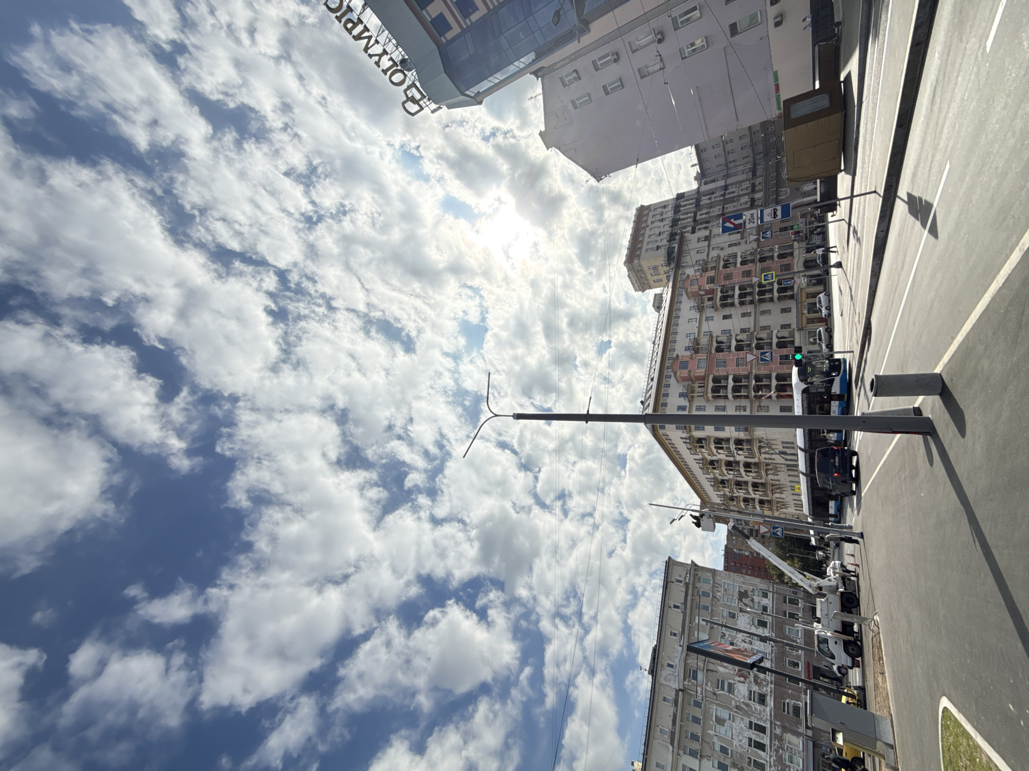 This is a street-level, eye-level, medium shot taken outdoors in Moscow, Russia, on a sunny day with scattered clouds. The perspective is looking upwards and slightly towards the right, capturing a wide view of a city street scene. In the foreground, a paved road with white lane markings curves gently to the left. A concrete planter is visible on the far right, casting a shadow.
Dominating the mid-ground and background are several multi-story buildings with architectural styles characteristic of Soviet-era residential and commercial structures. The building directly ahead and to the left is a weathered, light-colored structure with numerous windows. To its right, a taller building with a pale pink facade and balconies is prominently featured. Further to the right, a modern glass-fronted building with the sign "OLYMPIC" is visible at the top. Another light-colored building with many windows is positioned to the right of the pink building.
A variety of vehicles are present on the street. A white utility truck with a raised boom lift is parked on the left side of the road, possibly undergoing maintenance or repairs. A blue and white tram or bus is traveling on the right side of the road, and a black van is in front of it. Pedestrians are sparsely scattered throughout the scene, walking on sidewalks or near the road. Streetlights and utility poles are visible throughout the image, with overhead wires also present. The sky above is a bright blue with significant cloud cover, with sunlight filtering through some of the denser clouds. The overall impression is a dynamic urban environment under a partially cloudy sky.