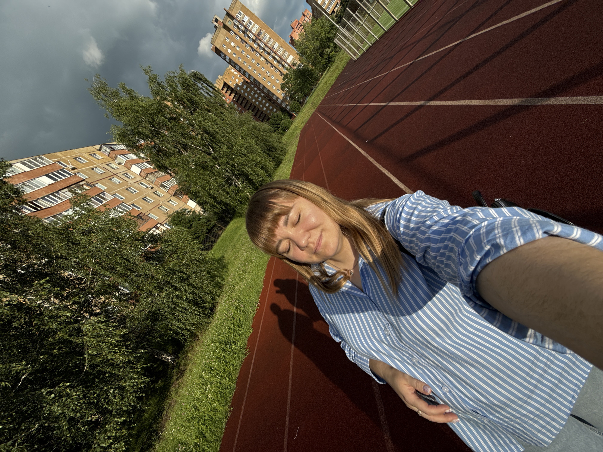 The media file captures a young woman with blonde hair and bangs, wearing a blue and white striped shirt, taking a selfie on a red running track. Her eyes are closed, and she has a peaceful expression, suggesting she is enjoying a moment of relaxation or repose. A faint smile plays on her lips. She is wearing a delicate necklace and a ring on her finger. To her left, a strip of lush green grass meets the edge of the track, with trees beyond. In the background, tall, multi-story buildings with brick facades and numerous windows are visible, interspersed with more trees. The sky is partly cloudy, with patches of dark clouds and lighter areas, indicating a dynamic weather condition, possibly a break in a storm or an approaching one. The sun is casting shadows on the track, suggesting it's daytime. The scene is set in an urban environment with residential buildings and a sports facility, specifically a running track. The overall impression is one of tranquil personal moment amidst an urban backdrop. No visible text is present in the image.