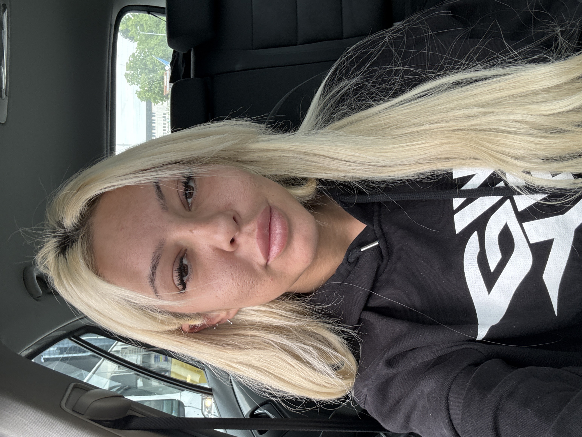 An angled, close-up selfie shows a young woman with long, light blonde hair and visible dark roots, looking directly at the camera. Her head is tilted, and her blonde hair cascades over her left shoulder and down the front of her black hoodie. She has fair skin, full lips, and dark eyelashes and eyebrows. Two small silver hoop earrings are visible in her left ear.
She is wearing a black hoodie with a large, stylized white graphic design on the front. Several long, thin strands of her blonde hair are visible on the dark fabric of the hoodie.
The setting appears to be inside a vehicle, likely a car. Dark car seats are visible in the background, and through the car windows, greenery (trees) and parts of urban buildings can be seen, suggesting an outdoor, daytime scene. The lighting is natural, consistent with daylight. The woman's expression is neutral and calm. The location context provided is Plovdiv, Bulgaria.